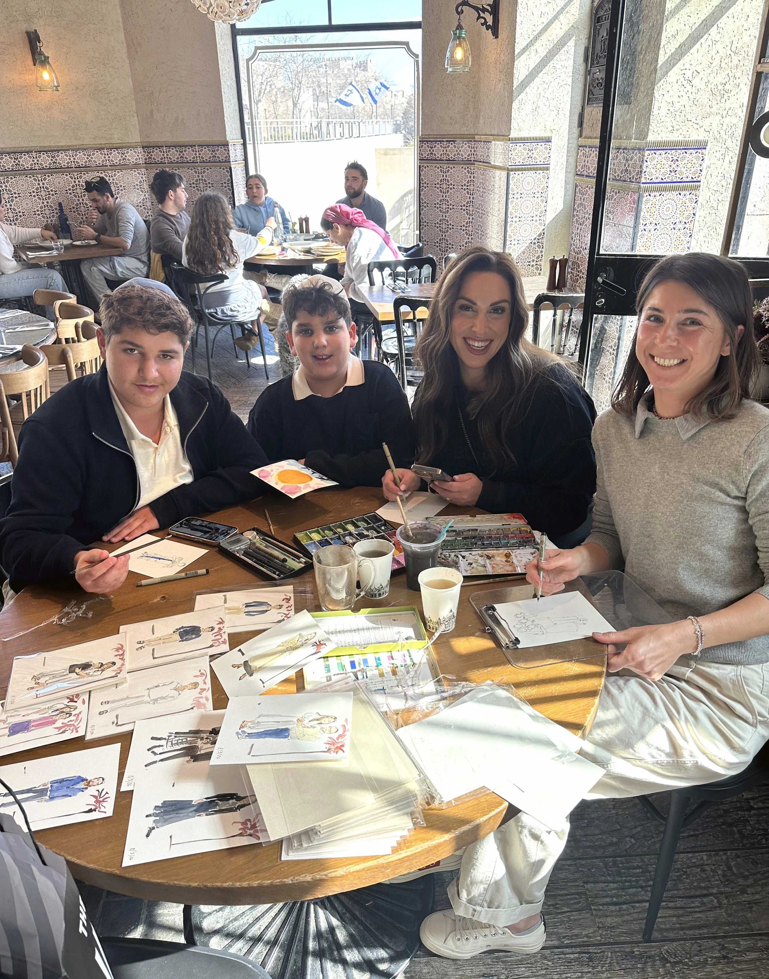 A group of four people sitting around a table in a restaurant, participating in an art activity. The table has fashion illustrations and watercolor paints, with two women and two boys smiling at the camera. Background shows other diners and large windows letting in daylight.