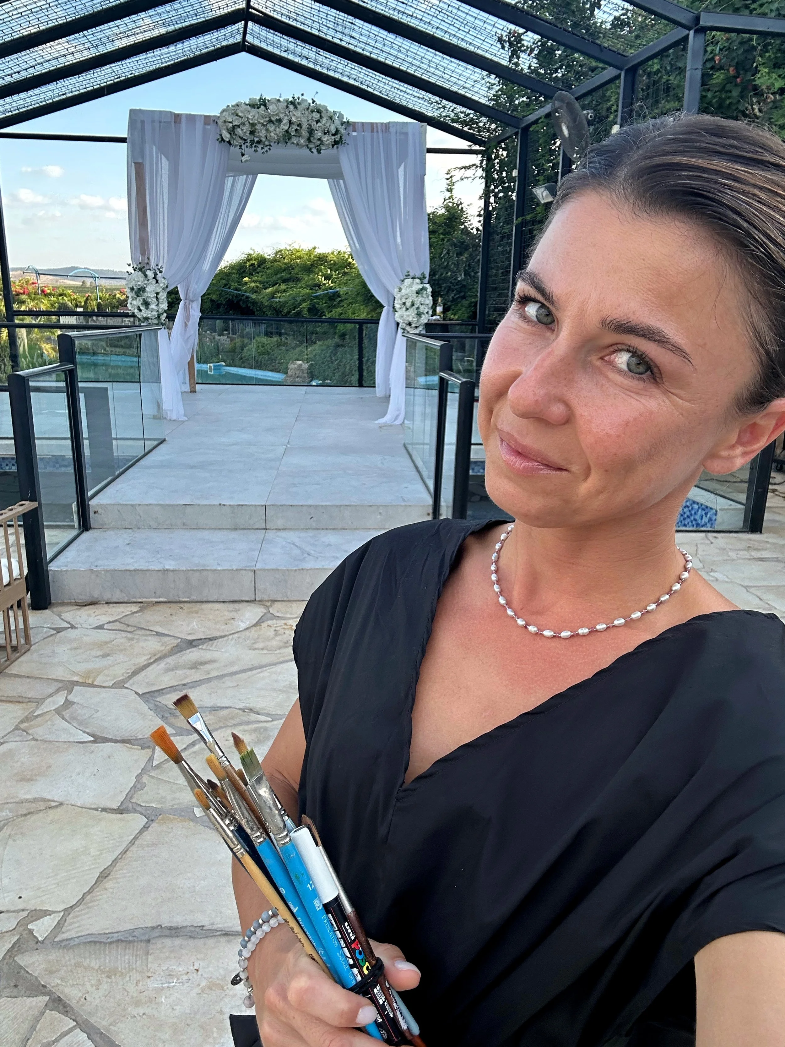 A woman holding paintbrushes on a stone patio, with a decorated outdoor wedding Chuppah in the background on a clear day.