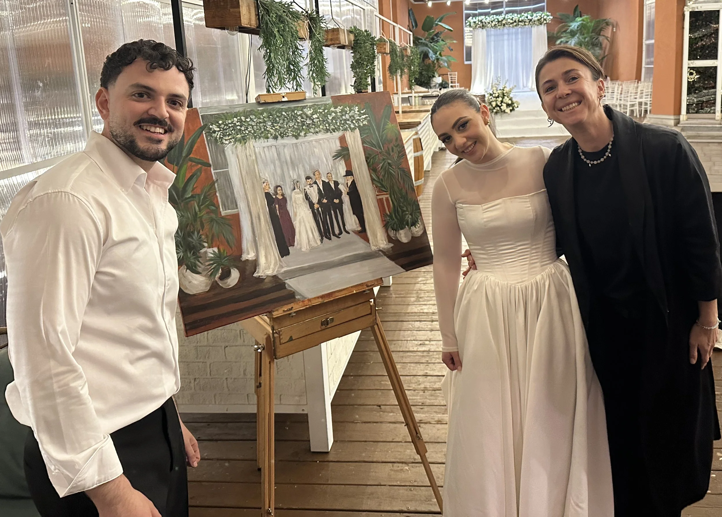 Three people standing next to a painting of a wedding scene. The couple in the wedding dress are smiling, with a woman and a man. The setting is indoors with wooden floors, plants, and large windows.