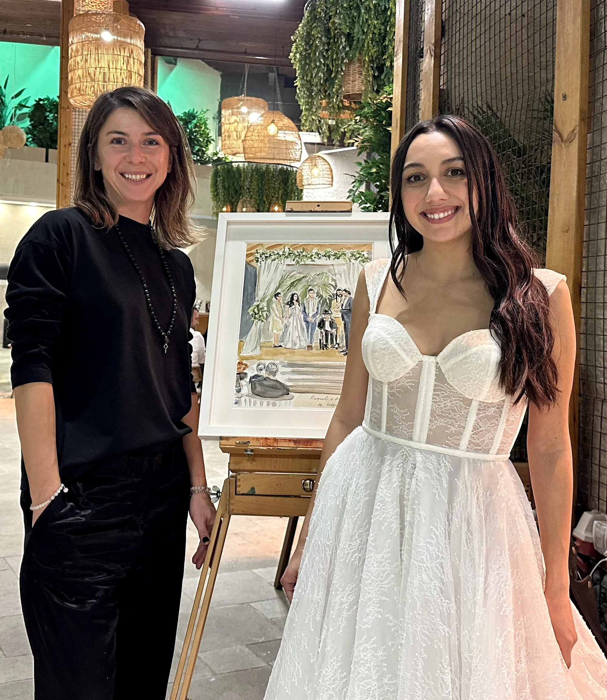 Two women standing in front of a framed painting at an indoor event, one in a black outfit and the other in a white lace dress.