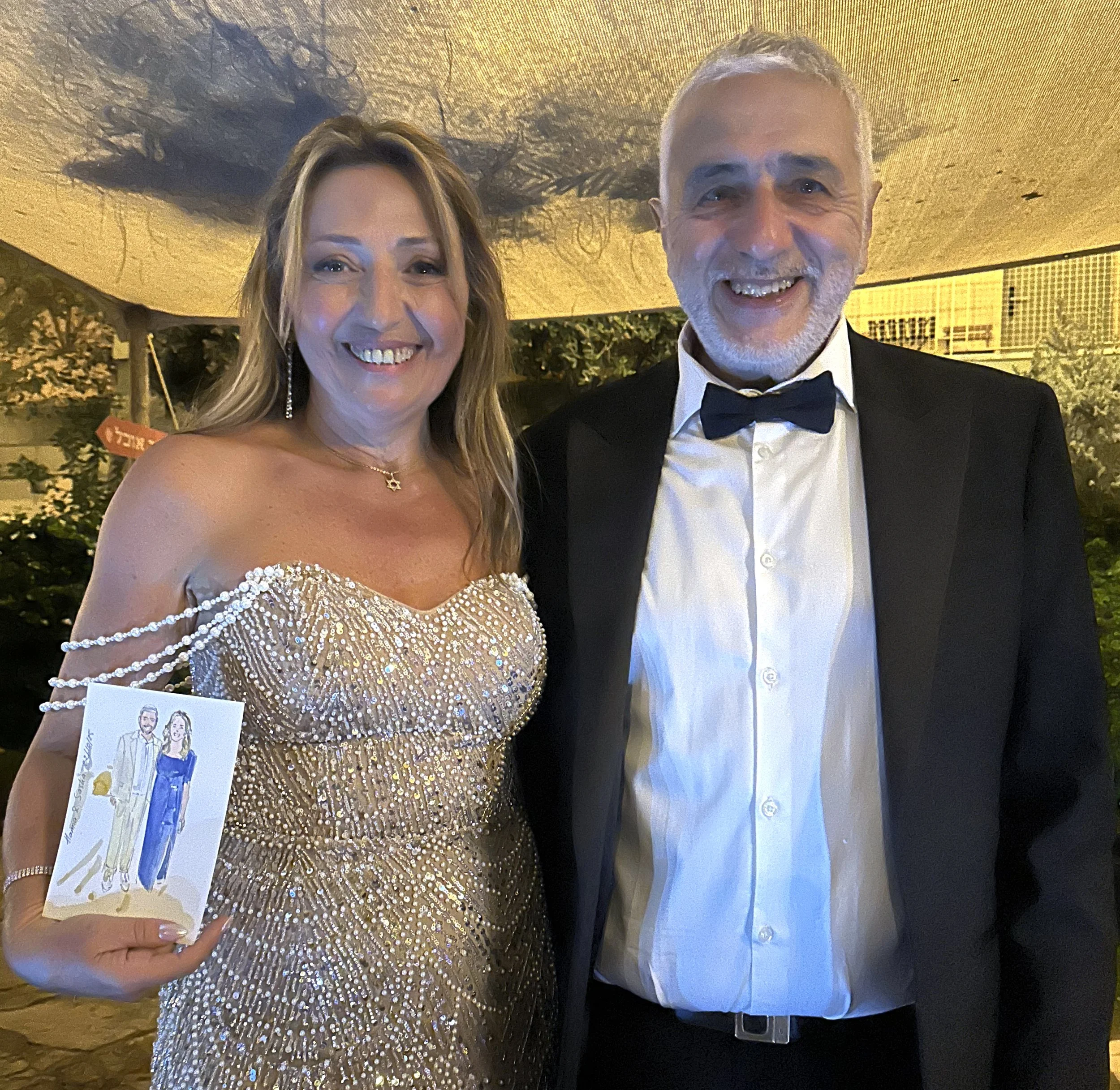 A smiling woman in a sparkly, beaded gold evening gown holding a card with a drawing of a man in a tuxedo and a woman in a gown, standing next to a smiling man in a black tuxedo and bow tie at an event.