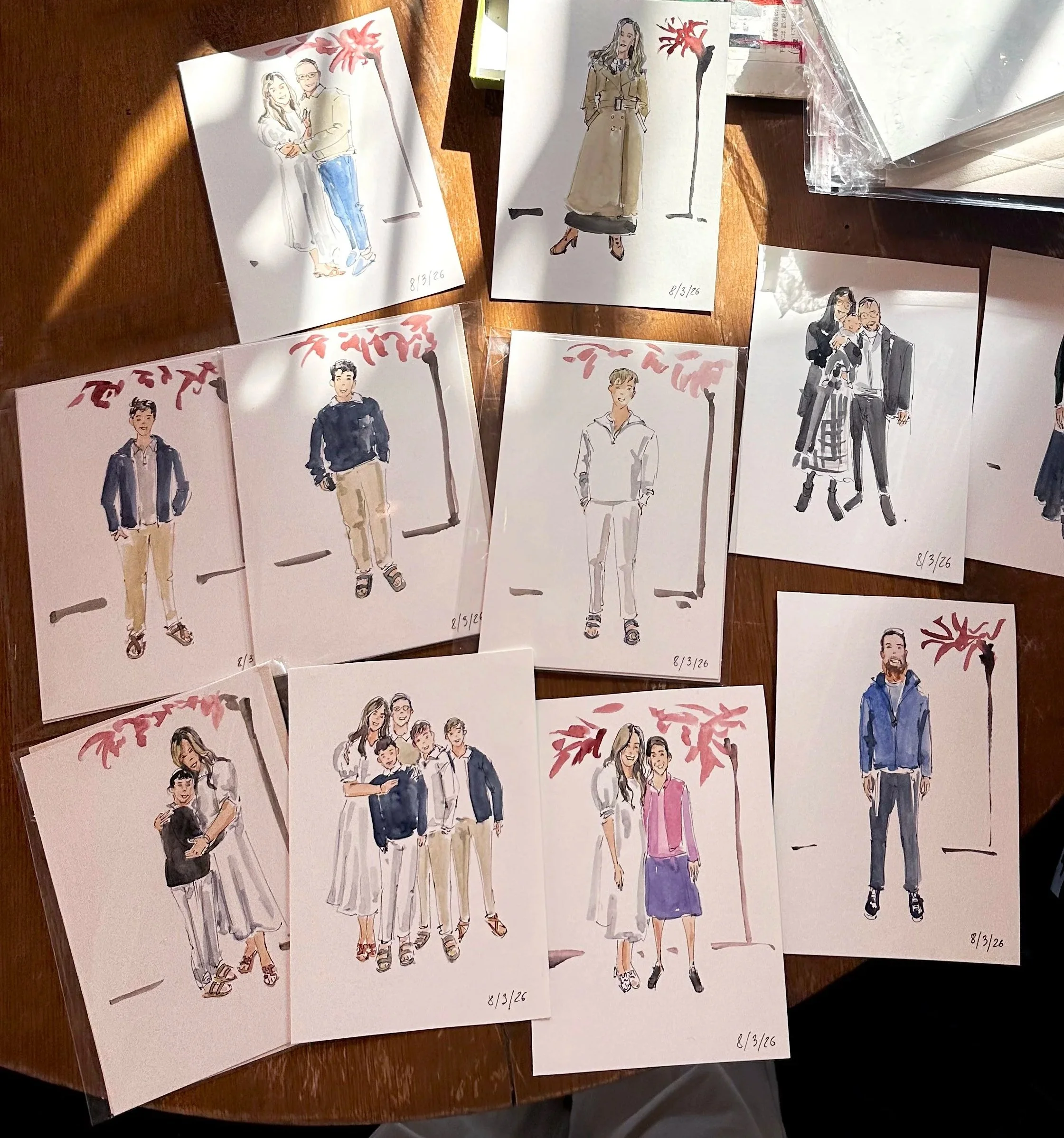 Collection of hand-drawn portraits and group sketches of people on white paper, arranged on a wooden table, with a small floral design and a date "8/3/16" on each.
