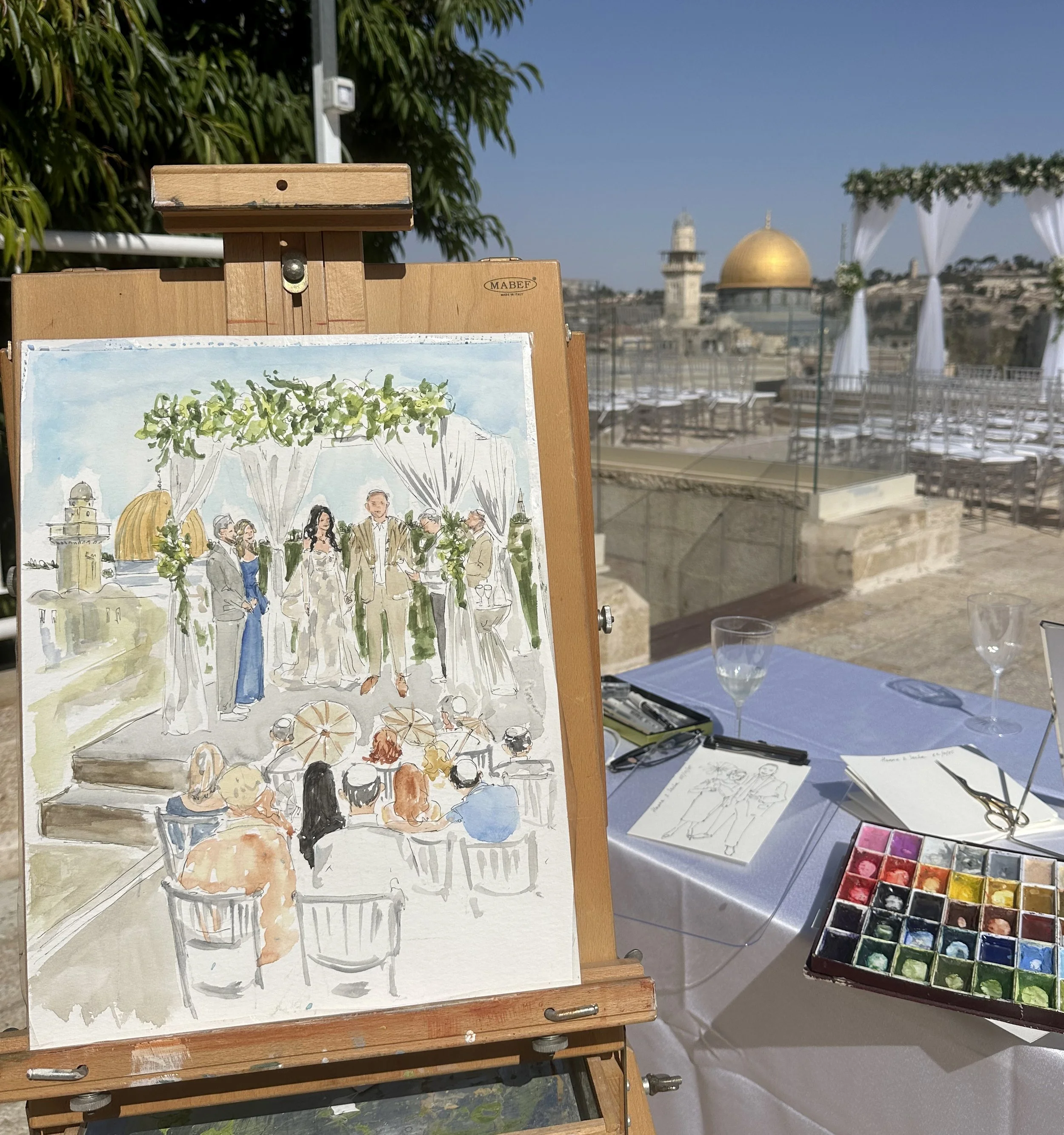 A watercolor painting of a wedding ceremony set up outdoors, with a bride and groom standing under a floral arch, surrounded by guests. The painting is on an easel, and there is a table with watercolor supplies and sketches in the foreground. The background shows a rooftop with a gold dome and a cityscape.