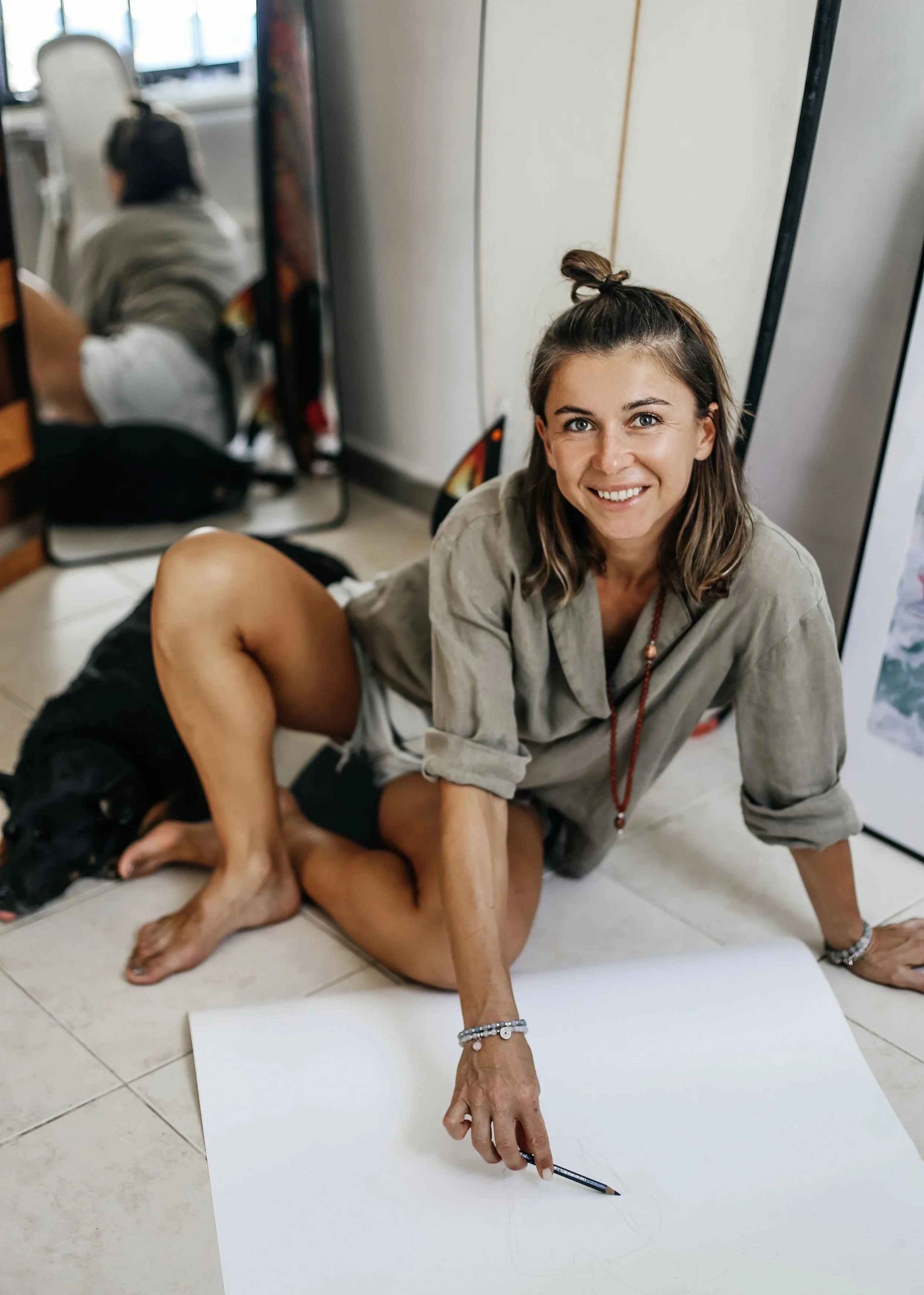 A woman artist, smiling, sitting on the floor of her studio next to a black dog, drawing on a large white sheet of paper with a pencil.