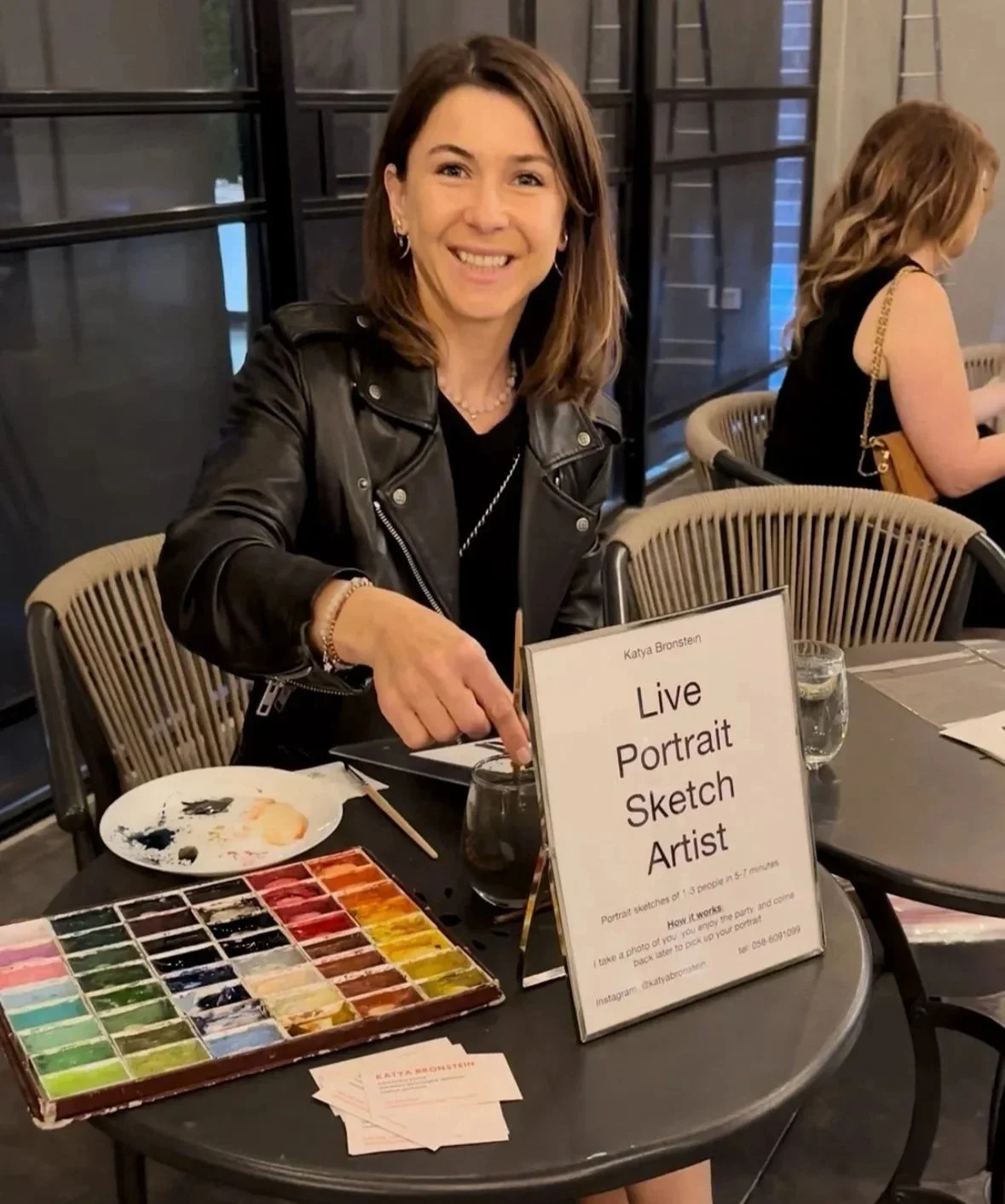 A woman sitting at a table with a sign that reads 'Live Portrait Sketch Artist' and a palette of watercolor paints, smiling at the camera