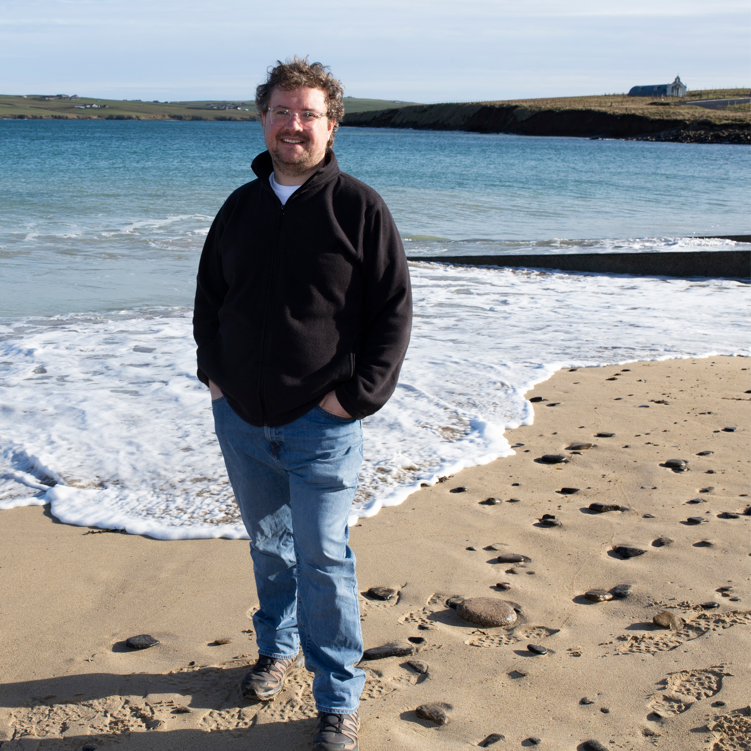 Kyle standing on a standing beach. Waves are lapping the shore. behind him and the Italian Chapel can be seen in the distance too