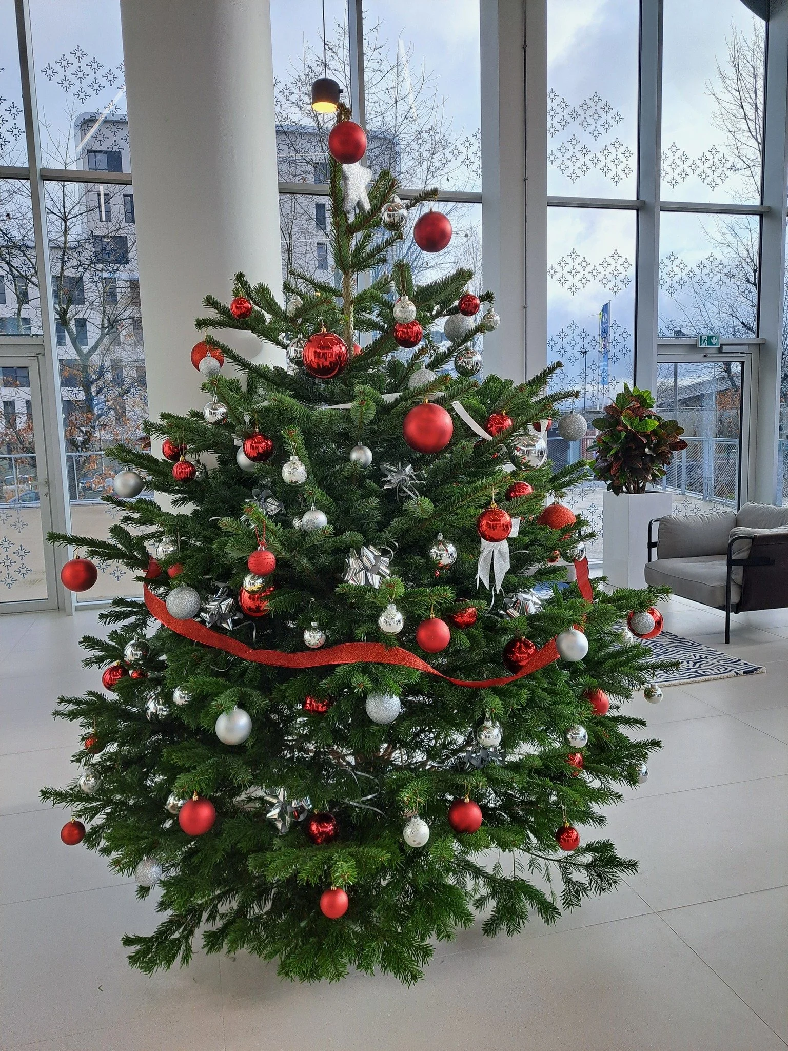 Un sapin de Noël décoré avec des boules rouges, argentées et des rubans rouges, placé dans une pièce moderne avec de grandes fenêtres et un décor hivernal visible à l'extérieur.