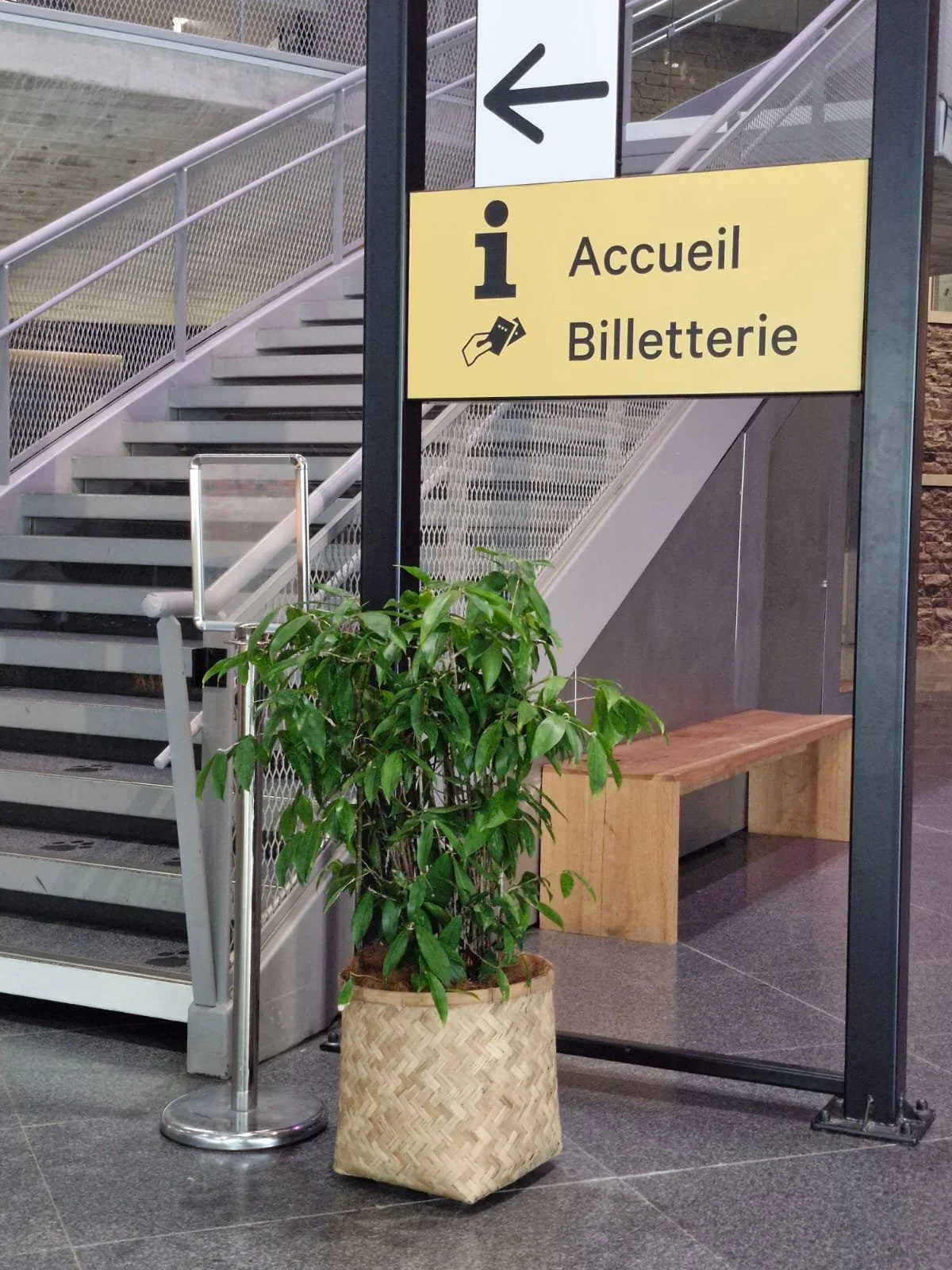 Une signalétique indiquant la direction vers l'accueil et la billetterie, avec un escalier en métal, une plante verte dans un panier en osier, un banc en bois, et un mur en pierre en arrière-plan.