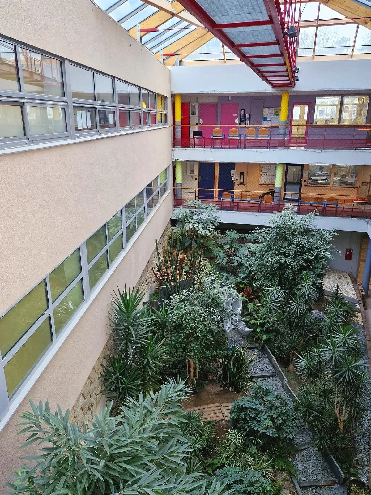 Halle intérieure d'un bâtiment avec un jardin central rempli de plantes variées et luxuriantes, ouverte au dessus par un plafond en verre, avec plusieurs étages visibles et des couloirs avec des sièges colorés.