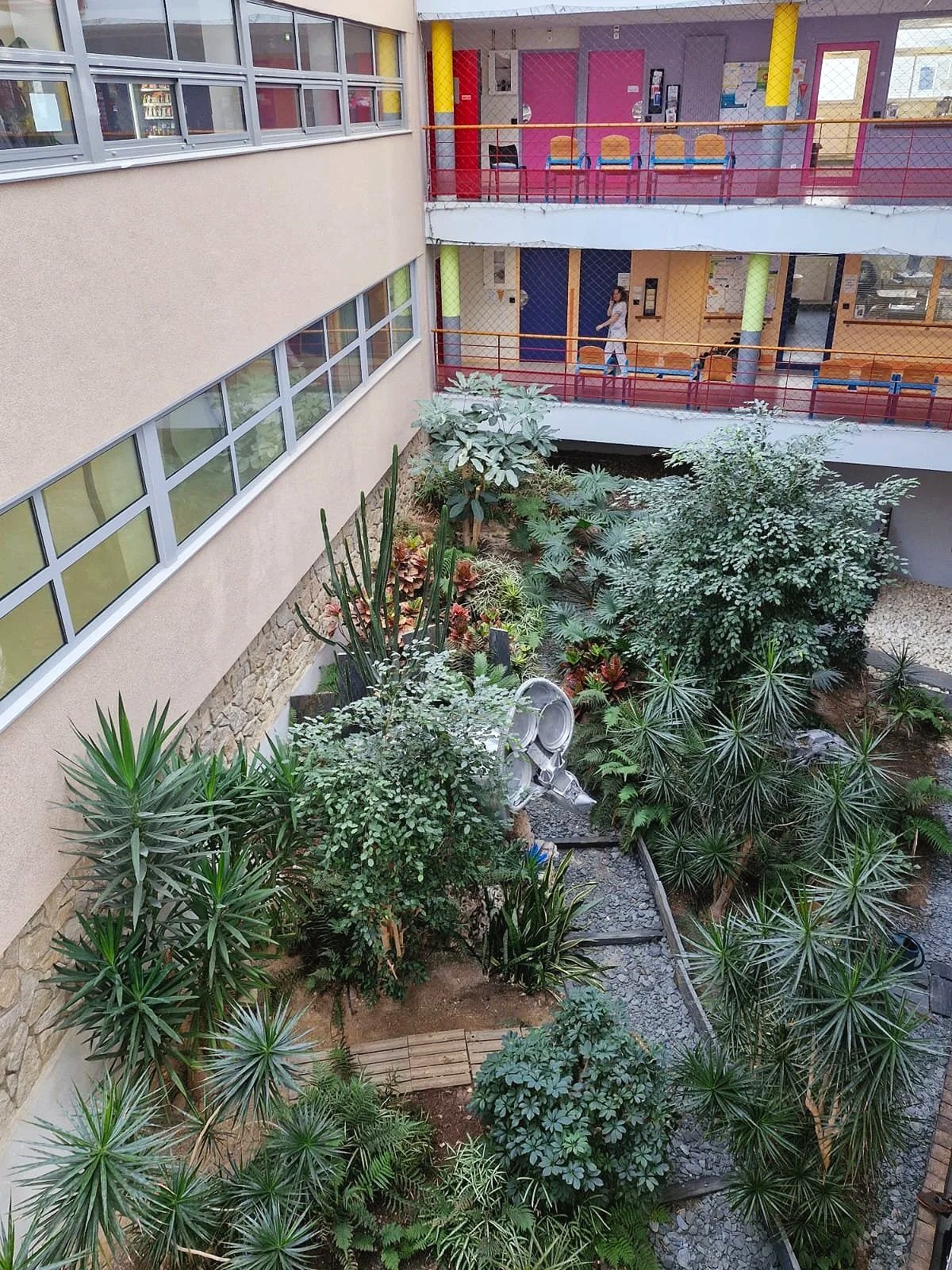 Vue d'un jardin intérieur avec diverses plantes vertes et un jardin de pierres, entouré de plusieurs étages avec des fenêtres et des couloirs colorés. Une personne marche dans le couloir supérieur.