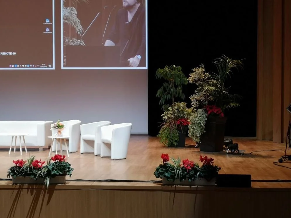 Une scène de conférence ou de présentation avec trois fauteuils et une table, décorée de plantes et de fleurs, sur une scène en bois, avec un grand écran à l'arrière.