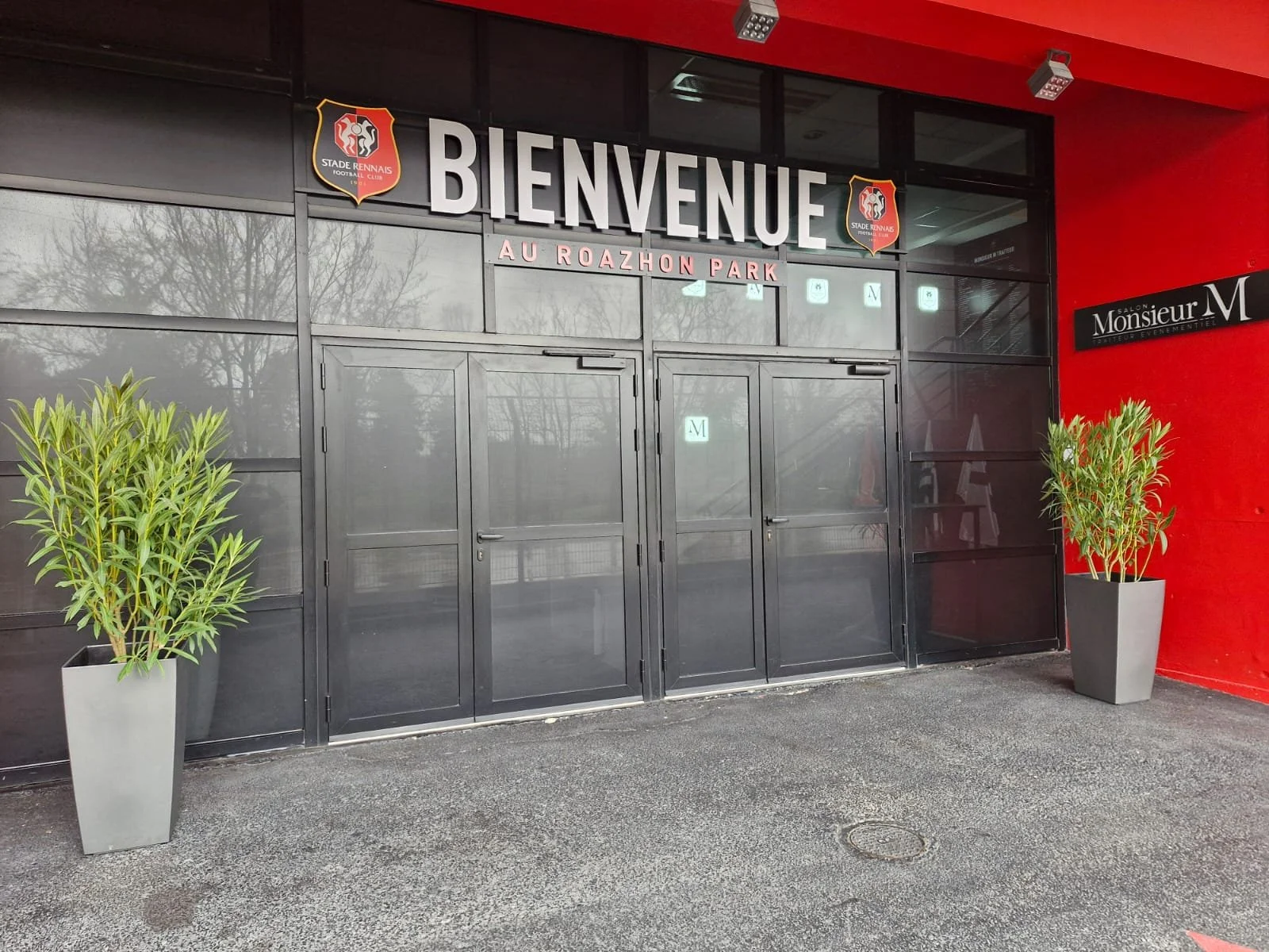 Entrée du stade Rennais Football Club avec deux plantes en pot de chaque côté, panneau de bienvenue 'BIENVENUE' au-dessus de la porte, et enseigne 'Salon Monsieur M' sur le mur à droite.