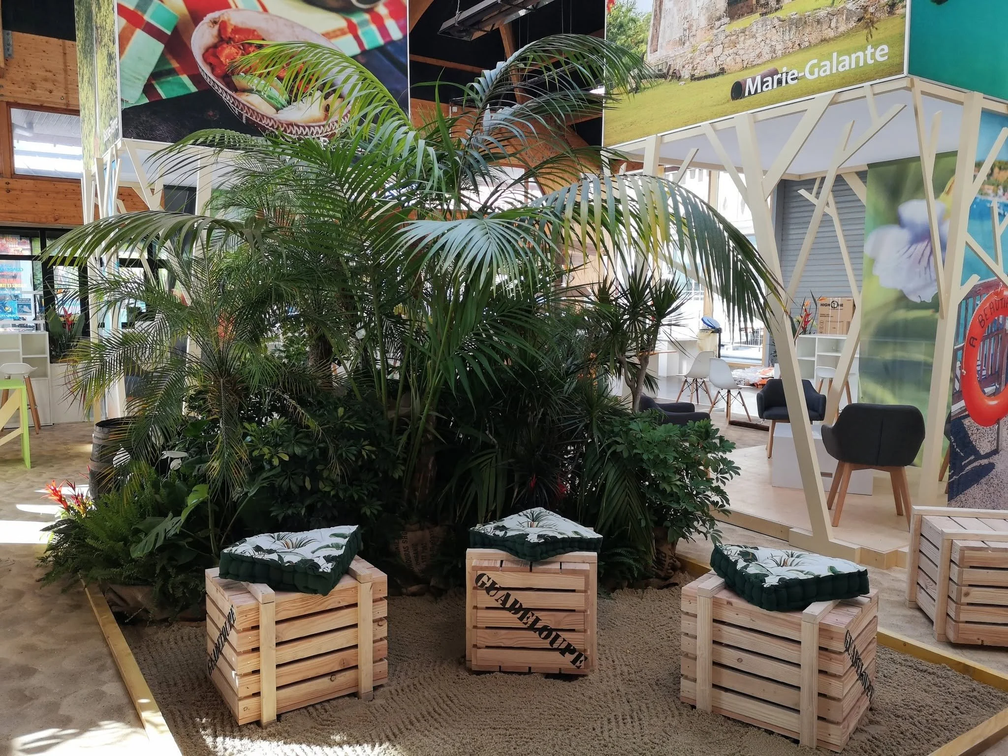 Intérieur d'un espace vert avec des plantes tropicales, des bancs en palettes en bois avec des coussins, et des affiches colorées suspendues au plafond.