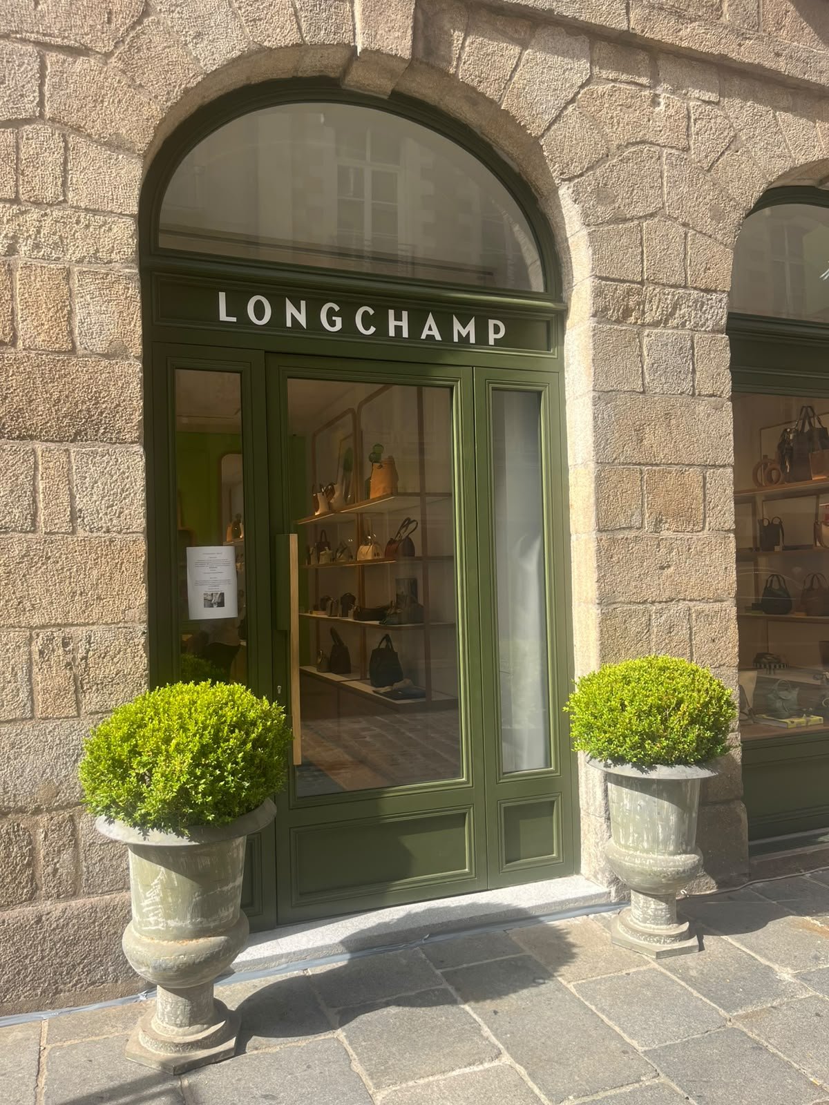 Vitrine d'une boutique de sacs à main avec le nom 'Longchamp' en lettres blanches sur une façade verte, flanquée de deux grands pots de fleurs avec des buissons verts, montée sur une façade en pierre.