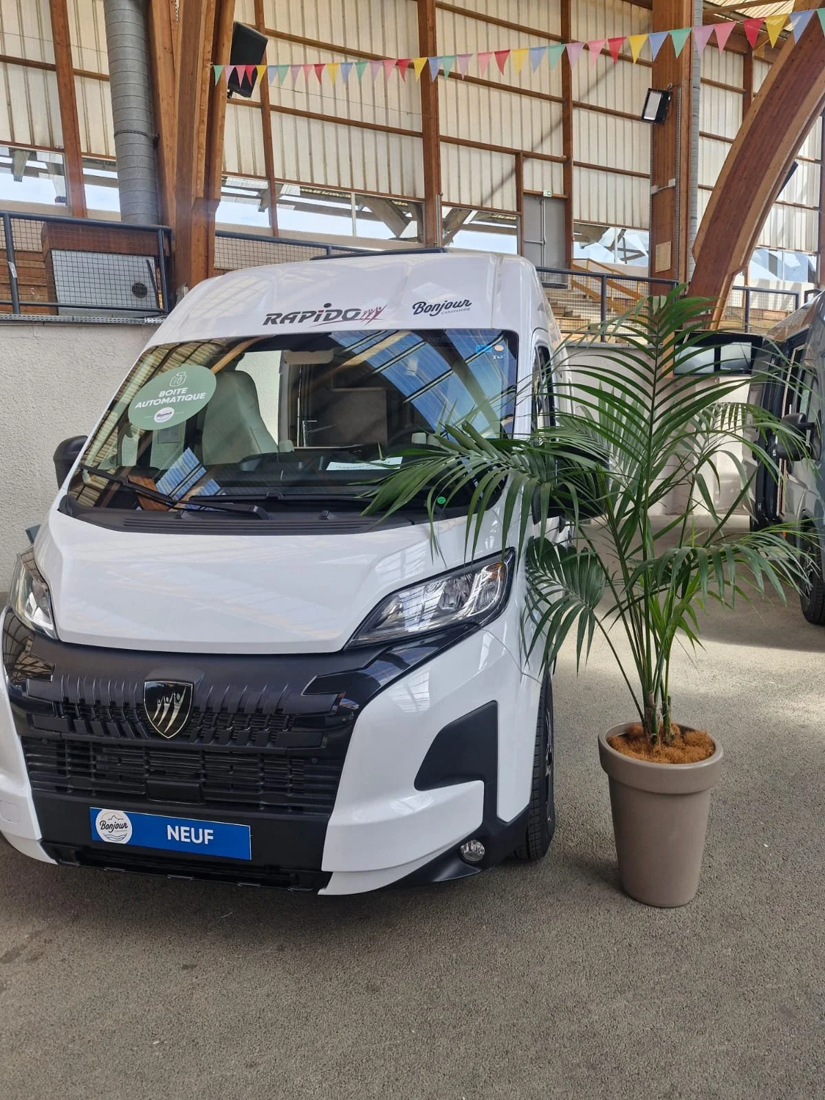 Voiture blanche neuve en showroom, avec un panneau indiquant "Boîte automatique" et un autocollant "Bonjour" sur le pare-brise, près d'une plante en pot, dans un bâtiment avec structure en bois et décorations de drapeaux colorés.