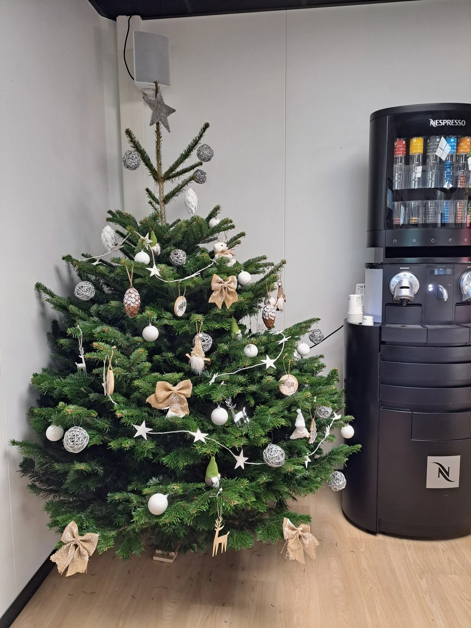 Un sapin de Noël décoré avec des boules blanches et argentées, des guirlandes, des nœuds en jute, et une étoile en haut. À proximité, une machine à café Nespresso.