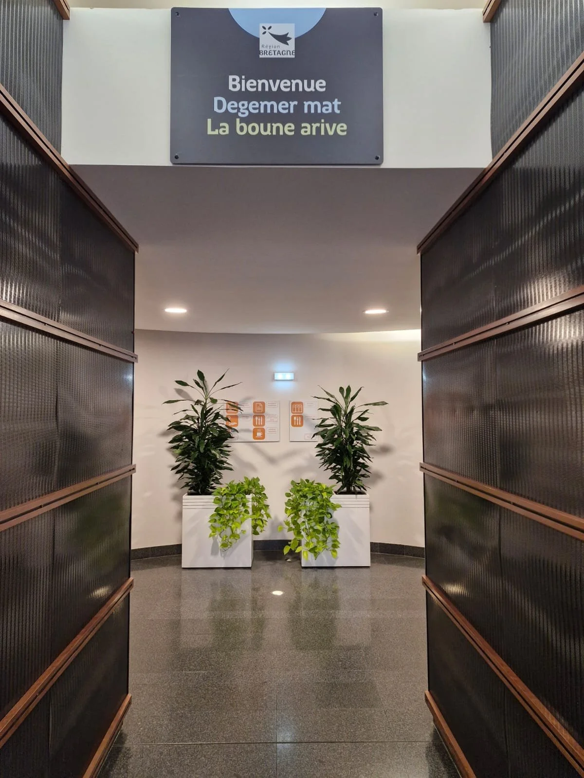 Entrée d'un bâtiment avec un panneau de bienvenue en français, deux grandes plantes vertes en pot blanc, et deux panneaux d'information à l'arrière sur un mur blanc.