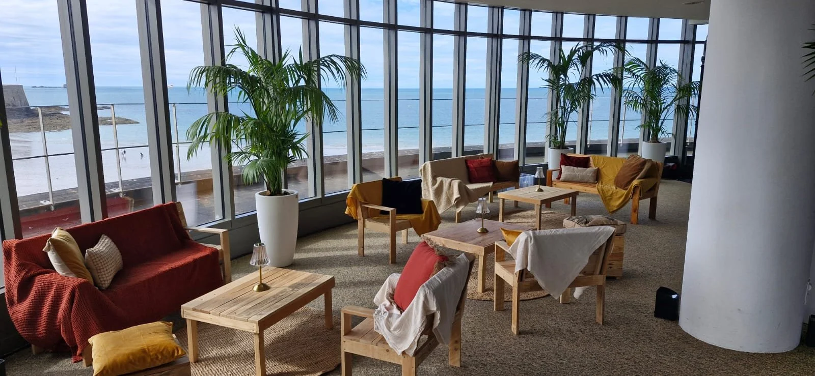 Salon avec grandes fenêtres offrant une vue sur la plage et l'océan. Mobilier en bois avec canapés, fauteuils, et tables, décoré avec des coussins et des lampes