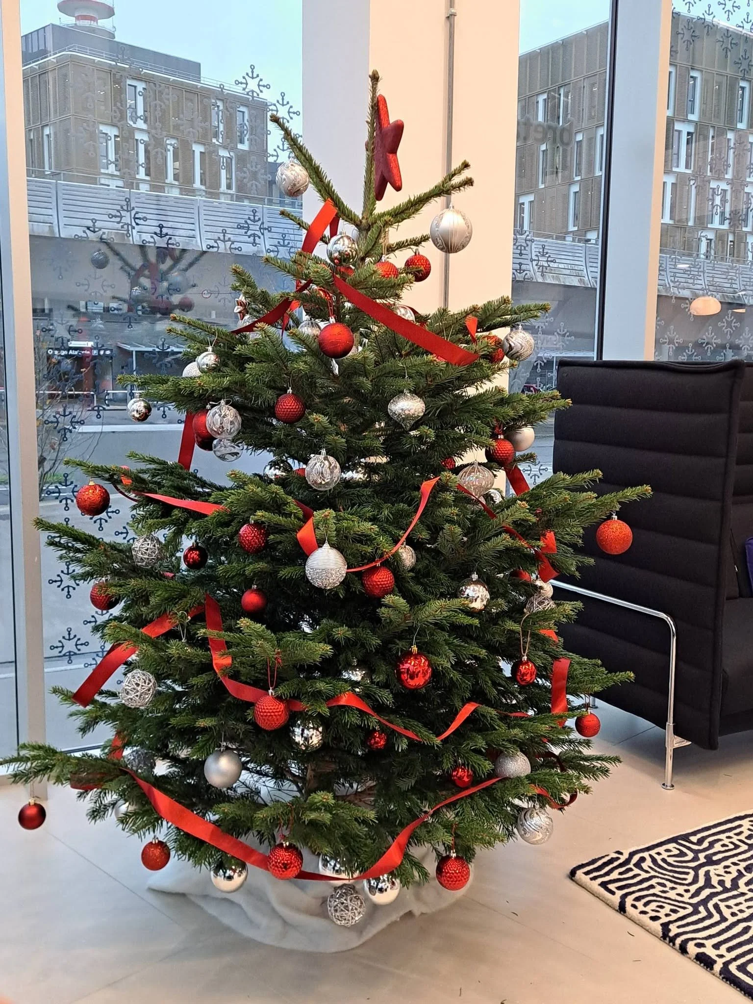 Un sapin de Noël décoré avec des boules rouges, argentées et blanches, ainsi que des rubans rouges, placé près d'une grande fenêtre dans une pièce moderne avec un fauteuil noir et un tapis noir et blanc.