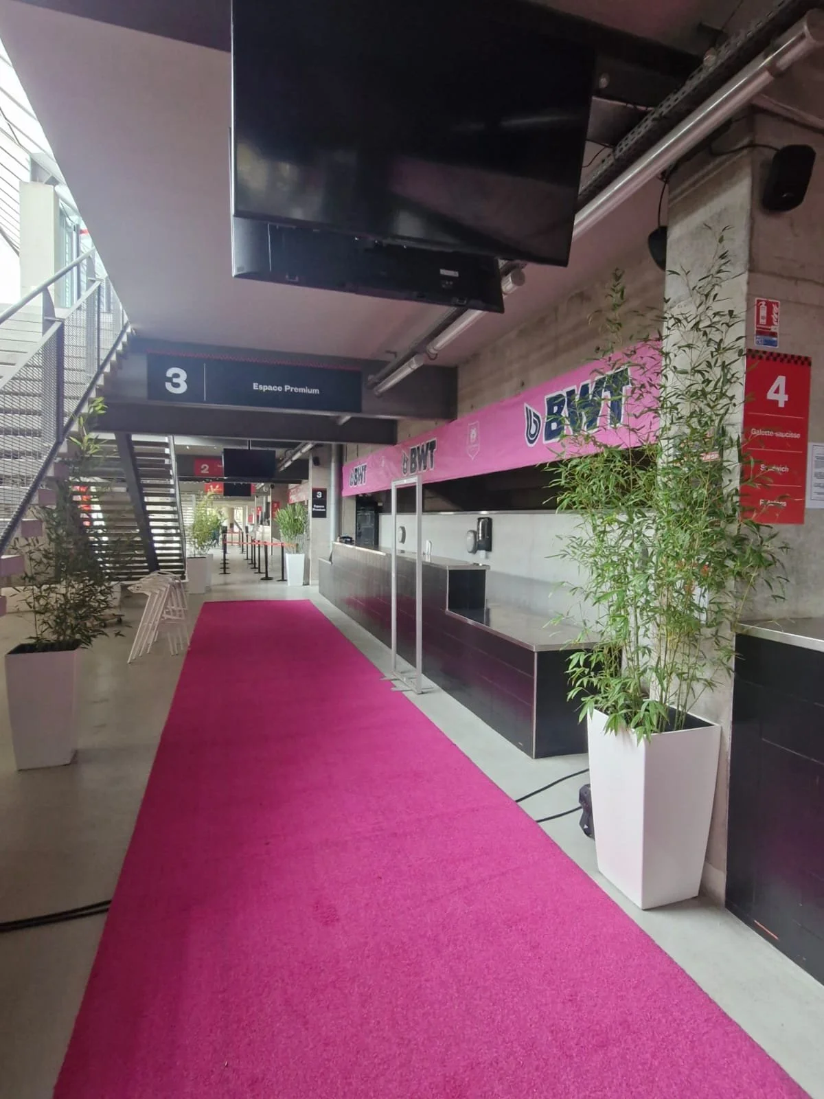 Intérieur d'un stade ou centre sportif avec un tapis rose, une billetterie, des plantes en pot, et des signalisations indiquant les entrées 3 et 4.