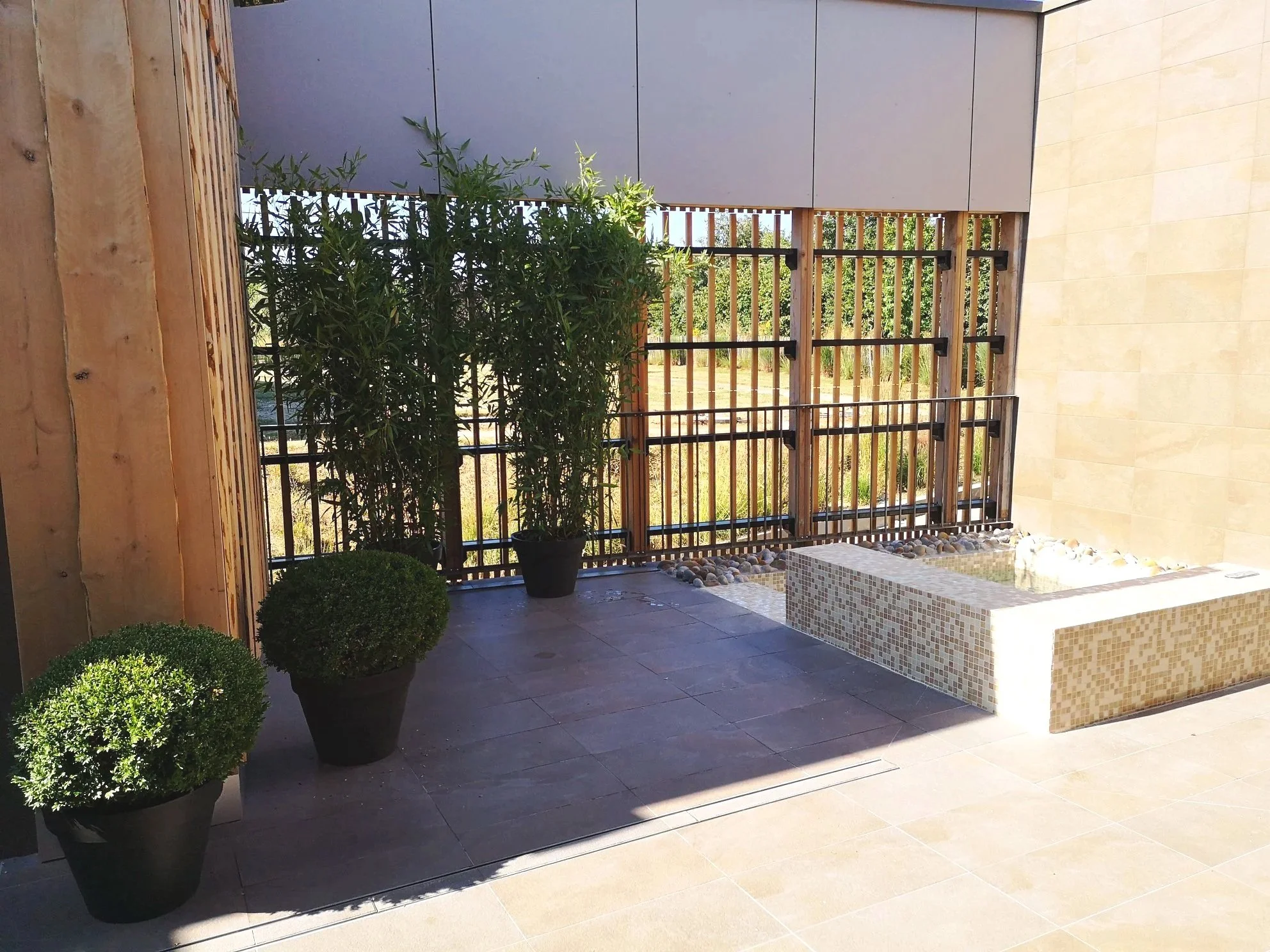 Un patio moderne avec un espace de jeu encadré par une clôture en bois et métal, des plantes en pot vertes et une petite fontaine ou bassin fait de mosaïque.