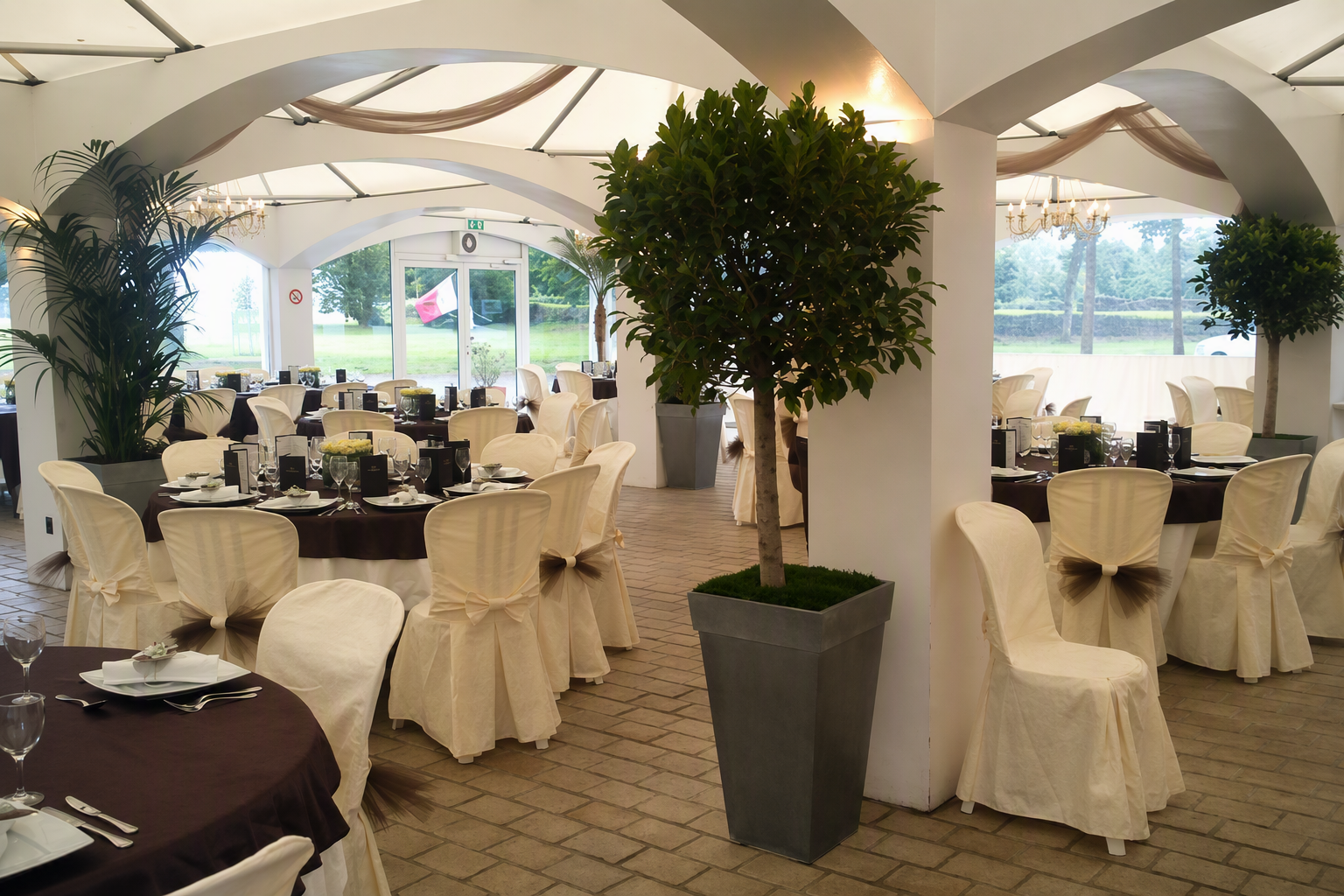 Salle de réception décorée pour un repas, avec tables rondes nappées de brun, chaises en tissu blanc avec noeud beige, grands pots de végétation, et vue sur un jardin extérieur à travers de grandes fenêtres.