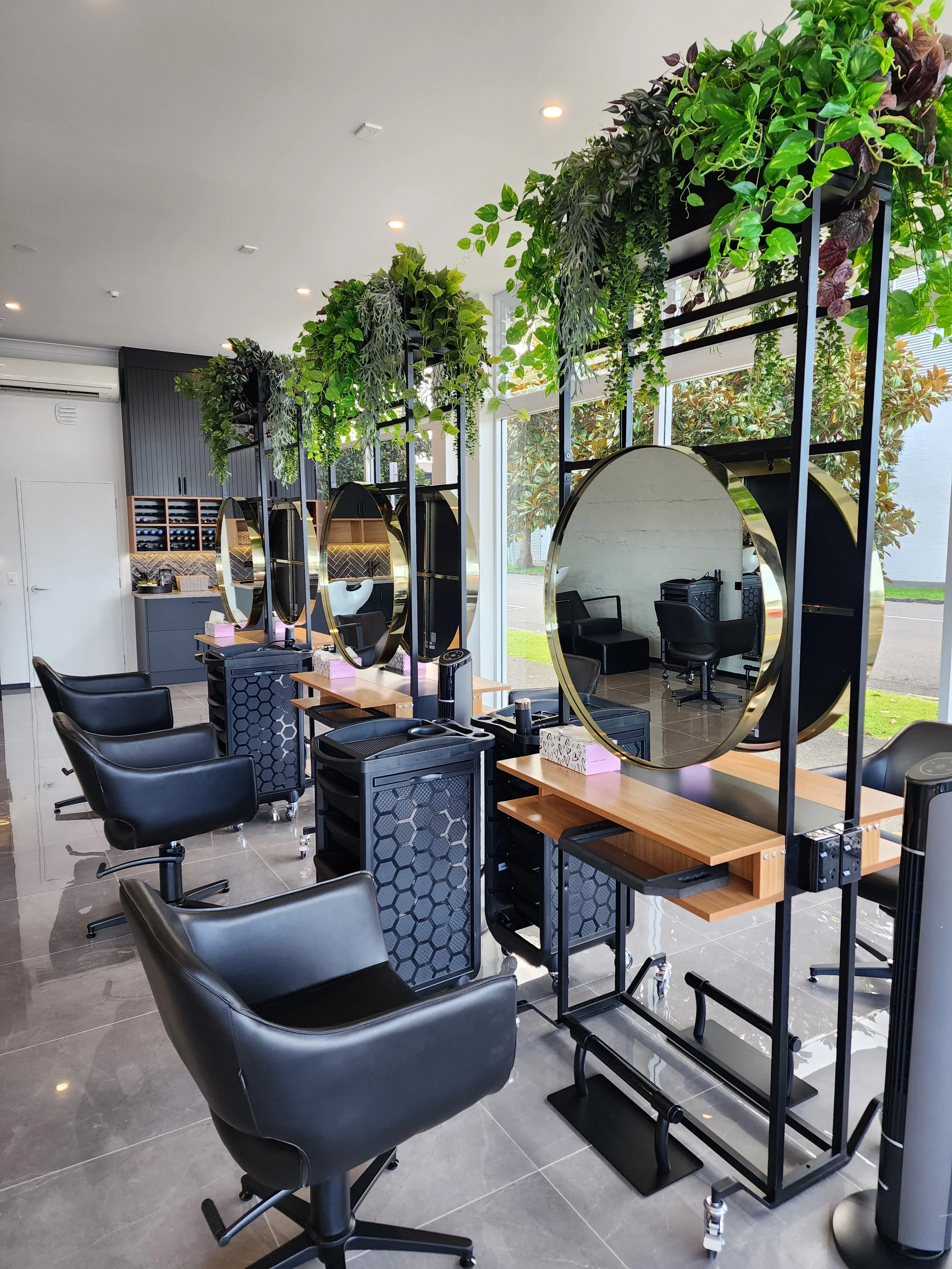 Salon de coiffure moderne avec plusieurs stations de coiffure, miroirs ronds avec bordures dorées, chaises noires, étagères noires avec des produits capillaires, décoration végétale suspendue au plafond, grande fenêtre naturelle illuminant l'espace.
