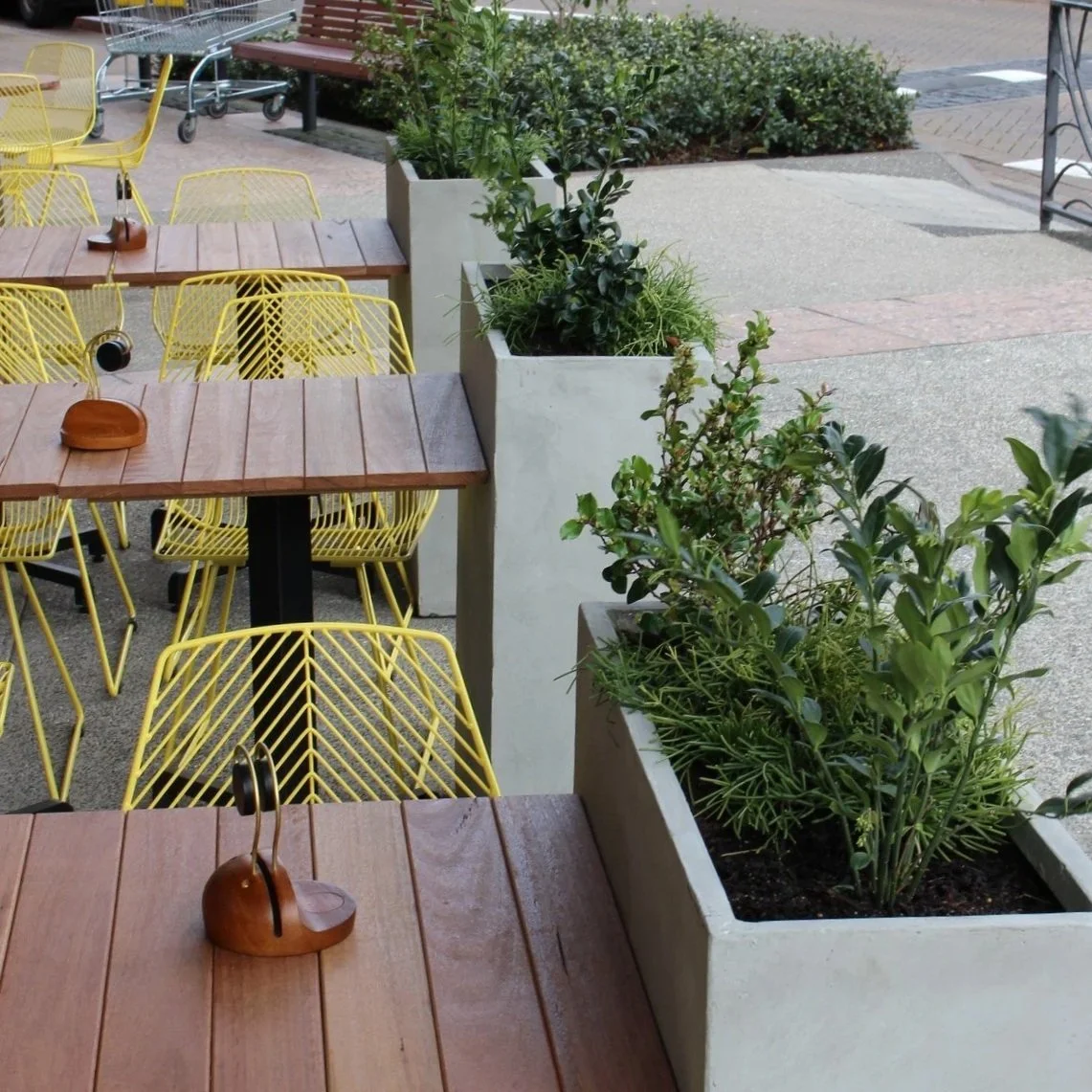 Terrace avec tables en bois, chaises jaunes, plantes en pot et vue sur la rue.
