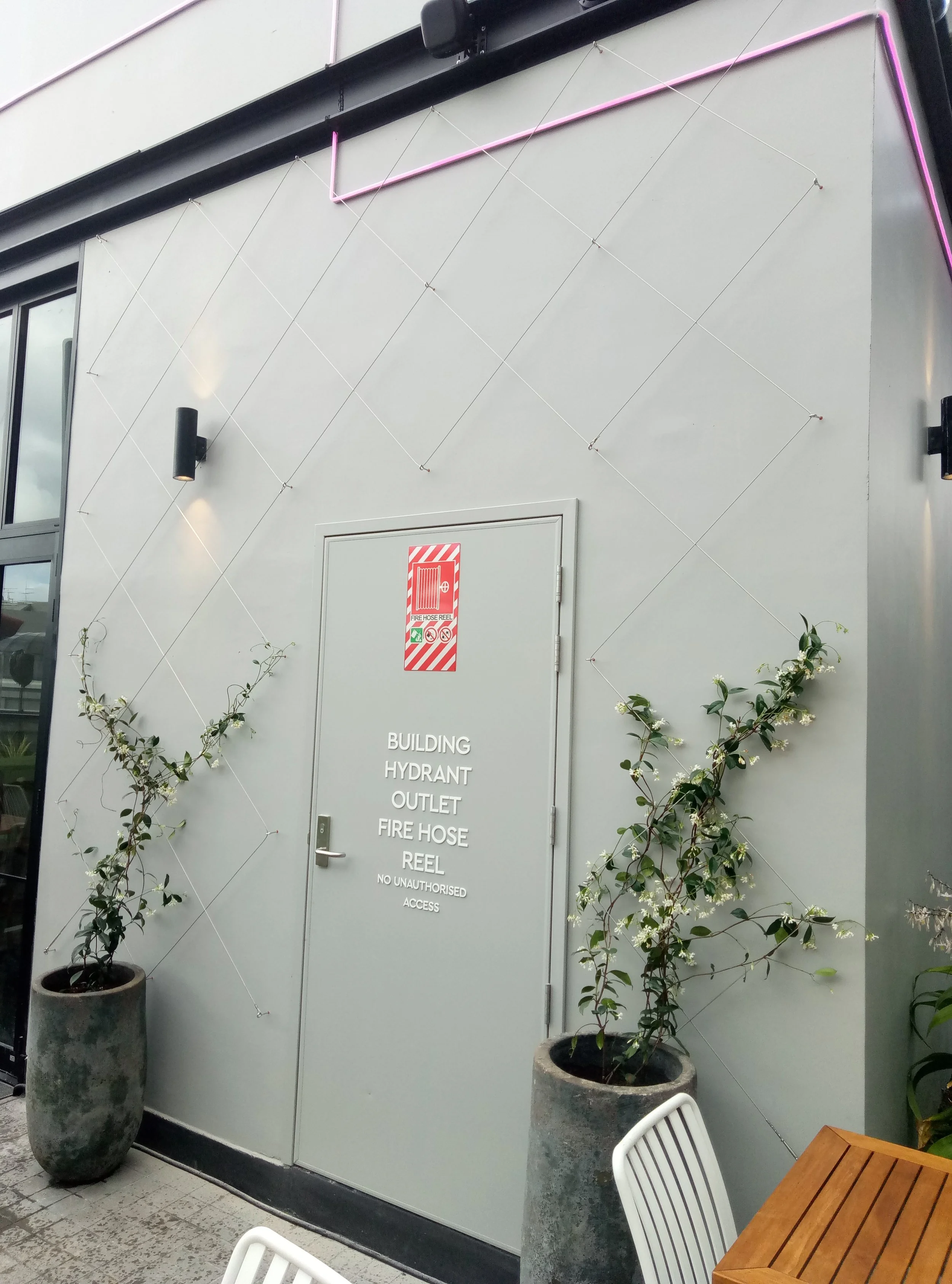Porte de sortie d'un bâtiment avec un panneau indiquant 'Building Hydrant Outlet, Fire Hose Reel, No Unauthorised Access'. Deux grands pots en béton avec des plantes à côté. Un panier en osier à droite et une table en bois. Deux chaises en plastique blanc, une à gauche et une à droite.