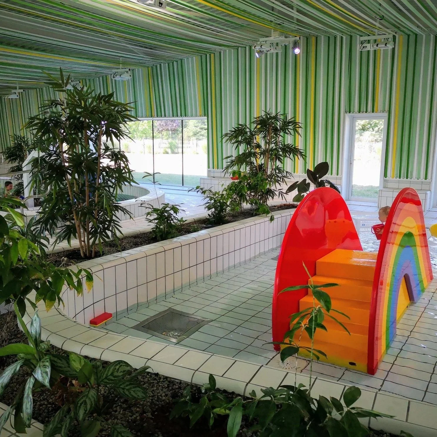 Intérieur d'une piscine avec des plantes, murs à rayures vertes et jaunes, et en arrière-plan une grande fenêtre laissant entrer la lumière naturelle. À droite, un jeune enfant à côté d'une structure en forme d'arc-en-ciel rouge, orange, jaune, vert, bleu et violet.