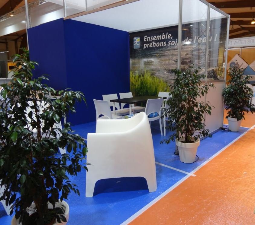 Espace d'exposition avec un coin salon, tables, chaises blanches, plantes en pots, mur bleu et panneau avec slogan en français.