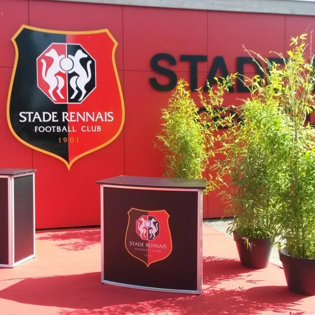 Logo du Stade Rennais Football Club avec des plantes vertes à côté, inscription partielle 'STADI' en arrière-plan.