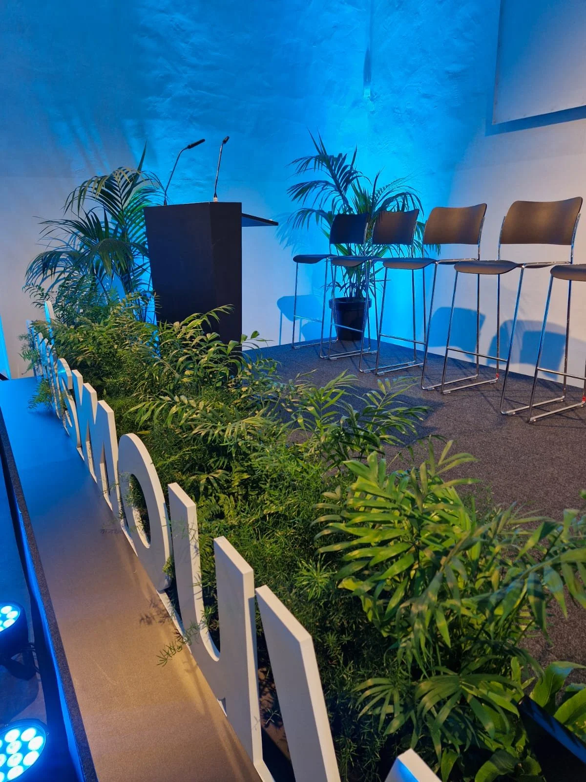 Une scène intérieure avec un podium noir, des chaises hautes en bois, des plantes vertes et un éclairage bleu, probablement une salle de conférence ou un espace pour un événement.