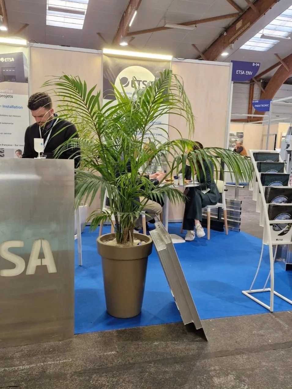 Un stand d'exposition avec une grande plante en pot au premier plan, plusieurs personnes assises et discutant, des panneaux d'exposition avec le logo ETSA, et des enseignes indiquant diverses sections de la foire dans un espace intérieur avec un plafond en bois et des lumières
