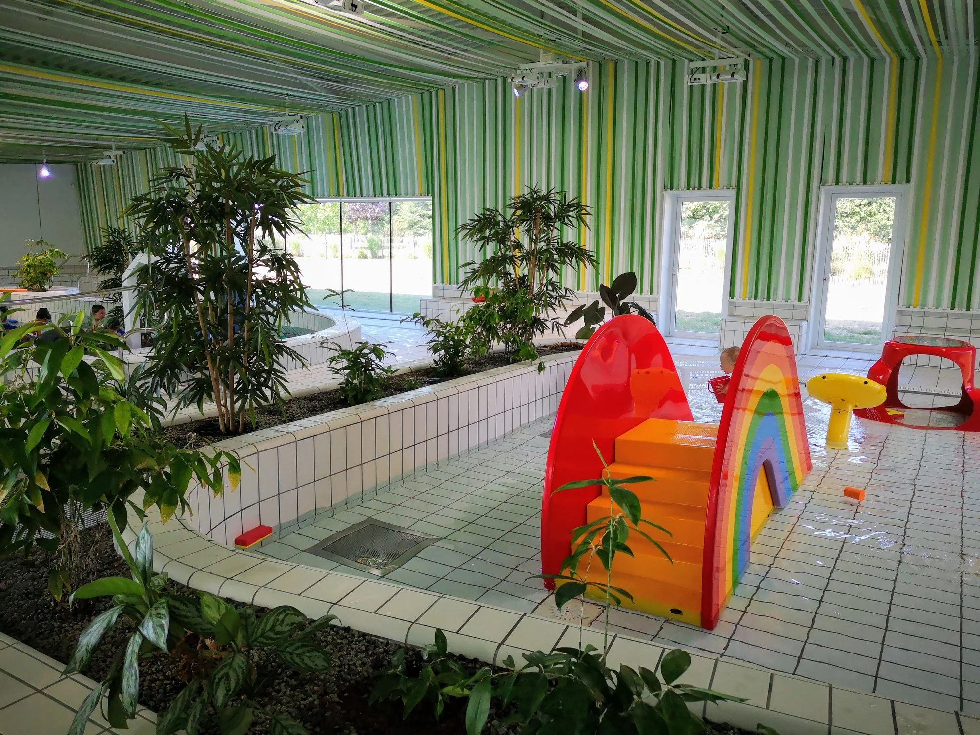 Une piscine intérieure avec des plantes vertes, des arbres en pot, des fenêtres laissant entrer la lumière naturelle, des jouets en plastique colorés, et un sol carrelé blanc avec des lignes vertes et jaunes sur le mur et le plafond.
