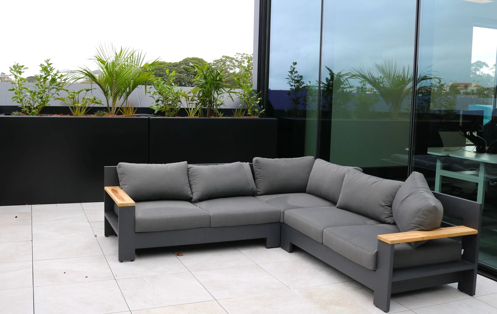 Balcon avec canapé gris, plantes en pot, et vue sur un paysage urbain à travers une grande fenêtre en verre
