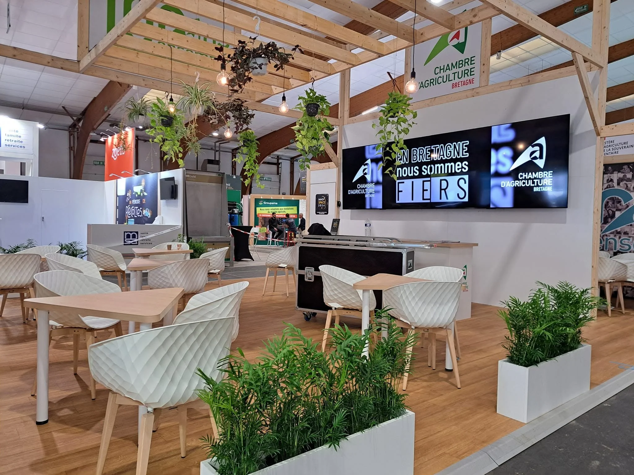 Un espace intérieur avec des tables en bois et des chaises blanches, décoré avec des plantes vertes en pots blancs, et un panneau d'affichage numérique qui mentionne la Bretagne et la chambre d'agriculture.