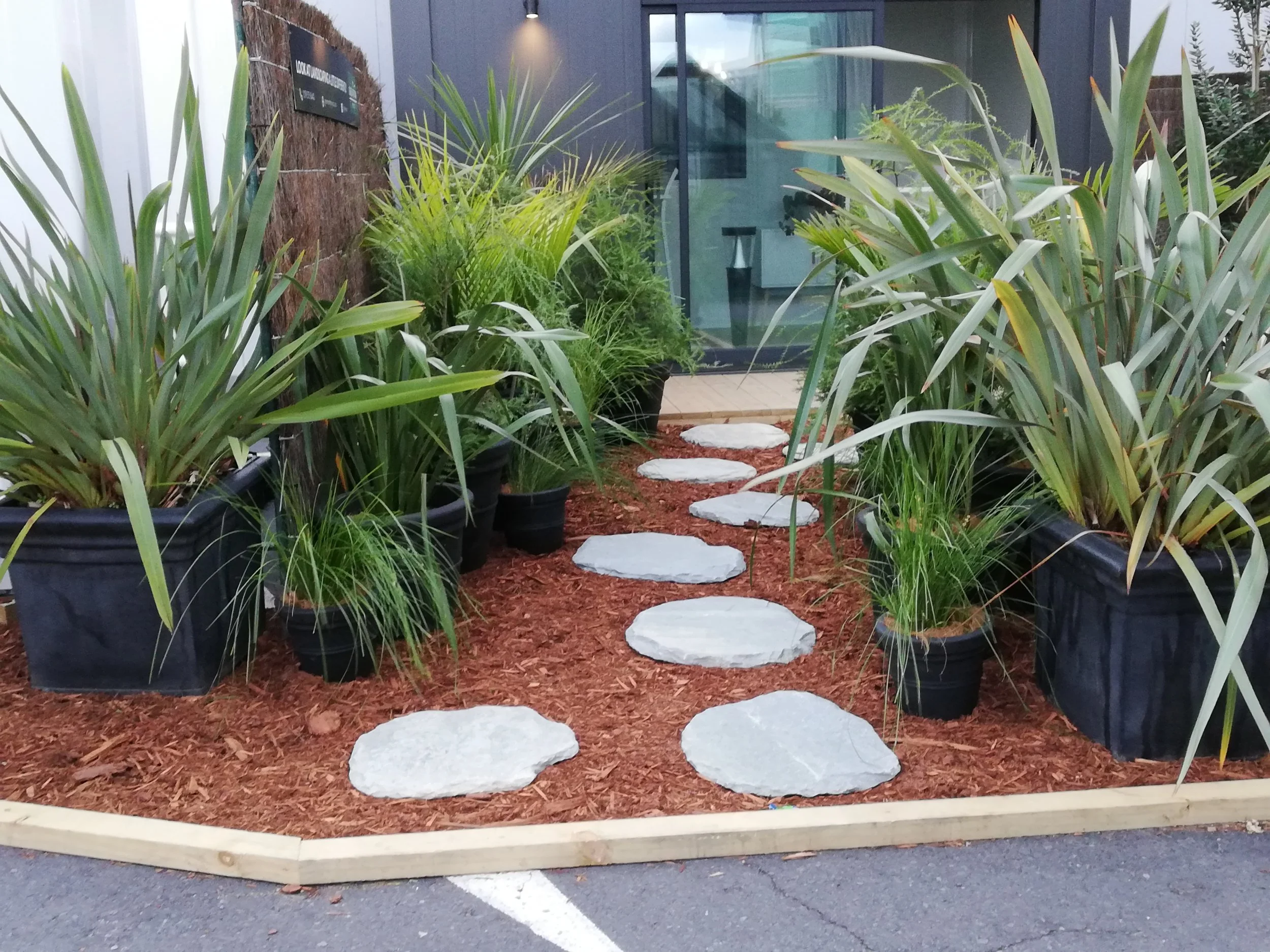 Jardin urbain avec des plantes vertes en pots noirs, un chemin de pierres, et un mur en bois derrière une clôture en bois, à l'extérieur près d'un bâtiment avec une porte en verre.