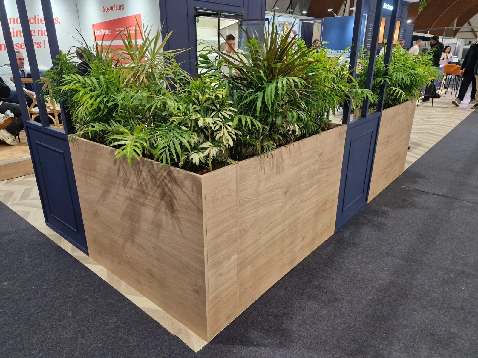Un mur de plantes vertes dans un grand cadre en bois placé dans un espace intérieur, avec des personnes assises et discutant en arrière-plan.