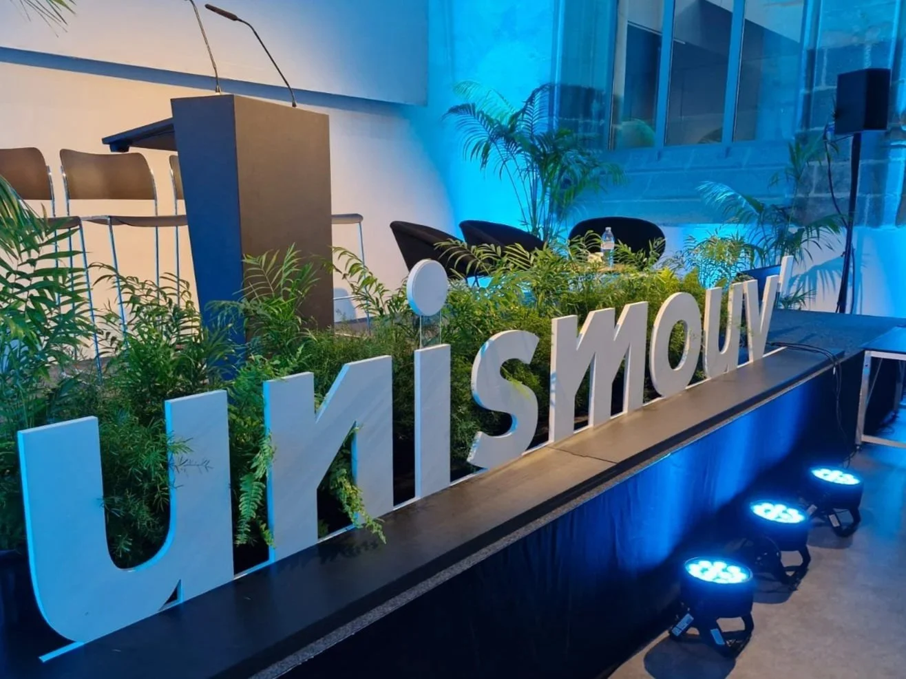 Décor intérieur avec plantes vertes, une bannière avec le mot 'UNISMOUV' en lettres blanches, une scène éclairée par des lumières bleues, un pupitre, des chaises et une enceinte audio.