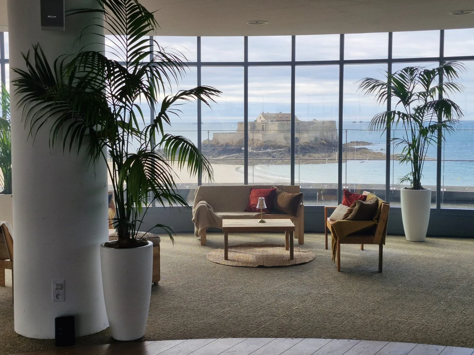 Salon avec vue sur la mer et un château à l'horizon, comprenant un canapé, deux fauteuils, une table basse et des plantes vertes en pot.