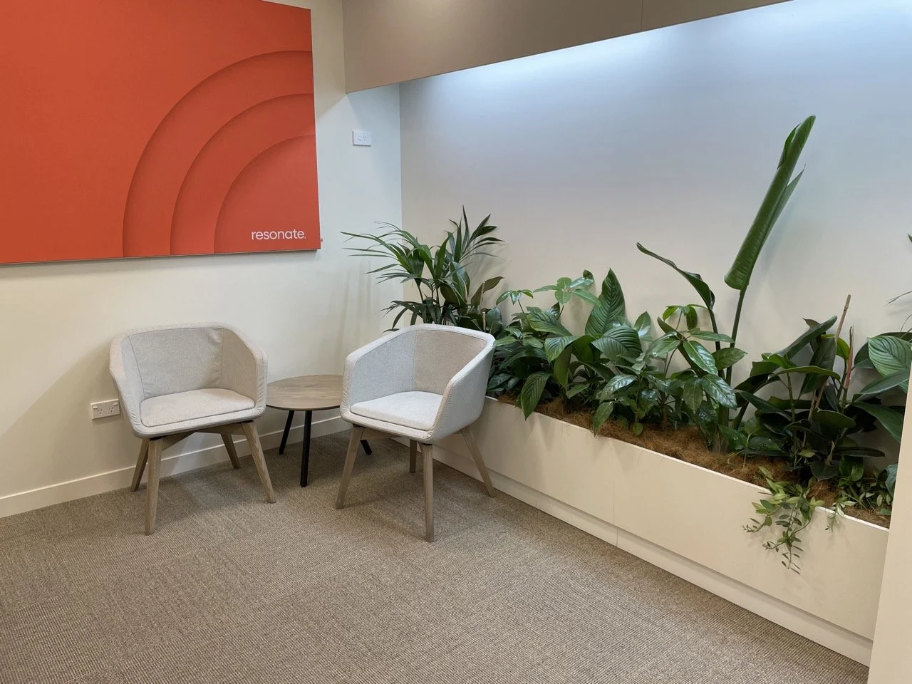 Espace d'attente avec deux fauteuils gris et une petite table en bois, entouré de plantes vertes dans une grande jardinière blanche, et un tableau orange avec le mot 'resonate' en bas à gauche.