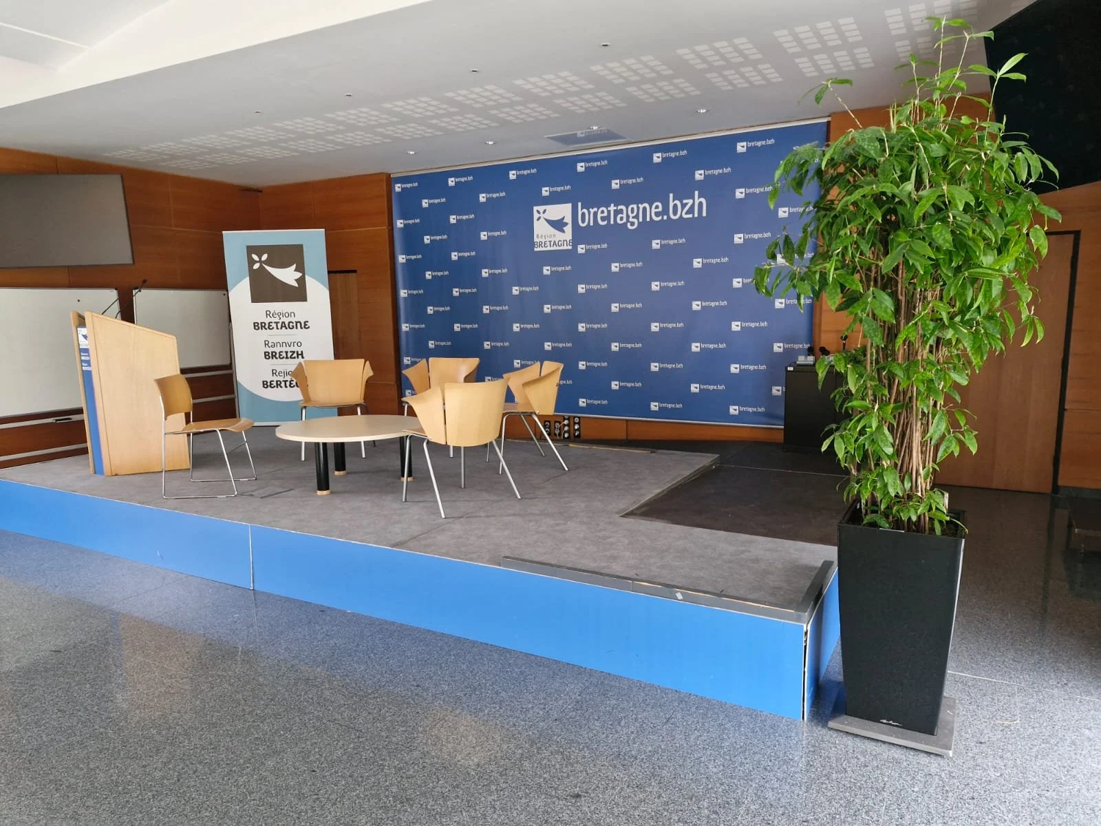 Une scène de conférence ou d'événement avec des chaises jaunes, une table blanche, une bannière avec le logo de la région Bretagne et un panneau d'affichage. Il y a aussi une grande plante verte dans un pot noir.