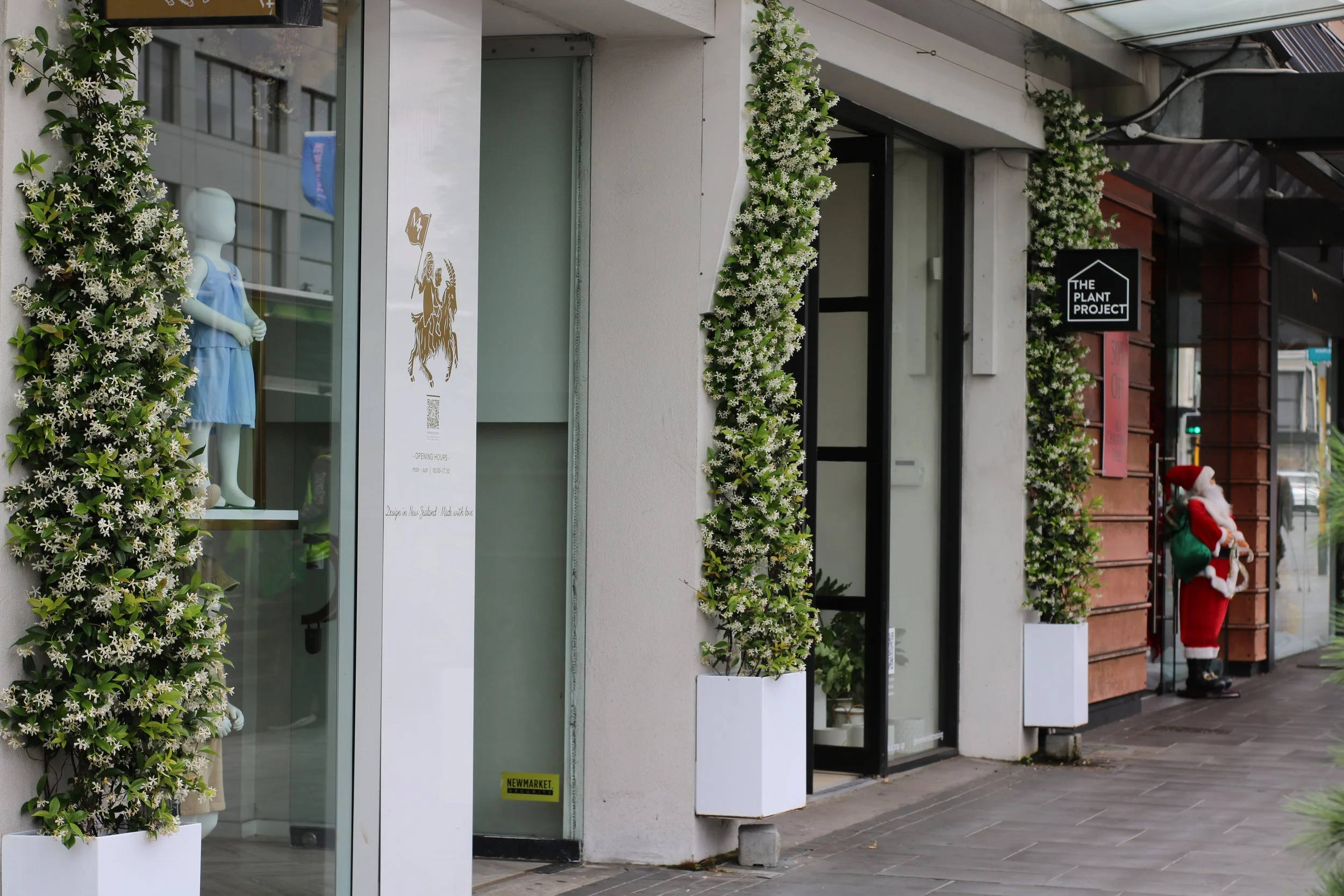 Façade d'une boutique avec des ornements de fleurs vertes et blanches, vitrine avec mannequin enfant portant une robe bleue, panneaux indiquant les heures d'ouverture et un logo "The Plant Project". Une décoration de Noël avec un Santa Claus est visible au coin de la rue.