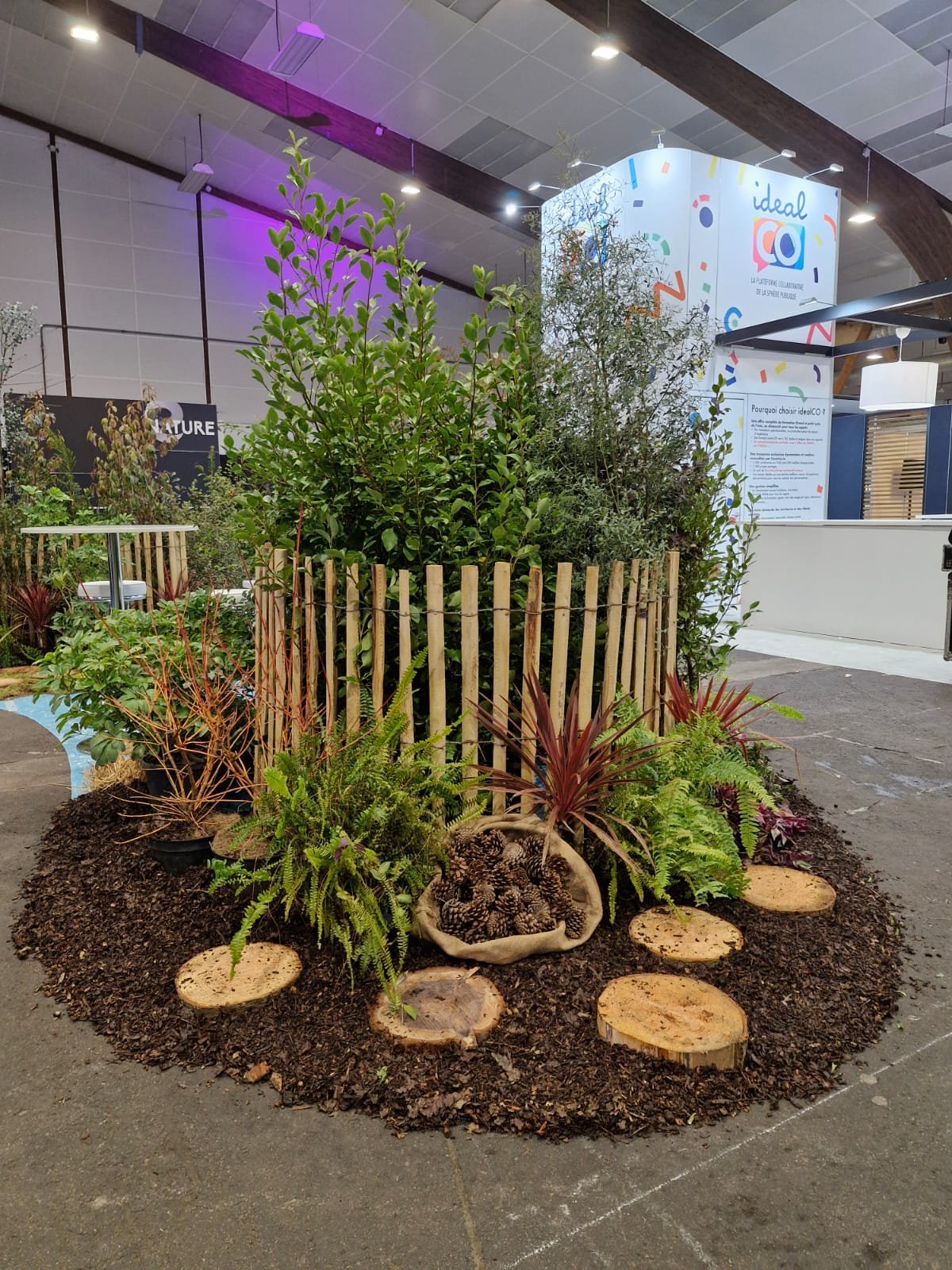 Un coin de jardin avec des plantes vertes et des arbustes entourés d'une barrière en bois, avec des chemins en rondins. Un mur de l'exposition Idealc est visible en arrière-plan.