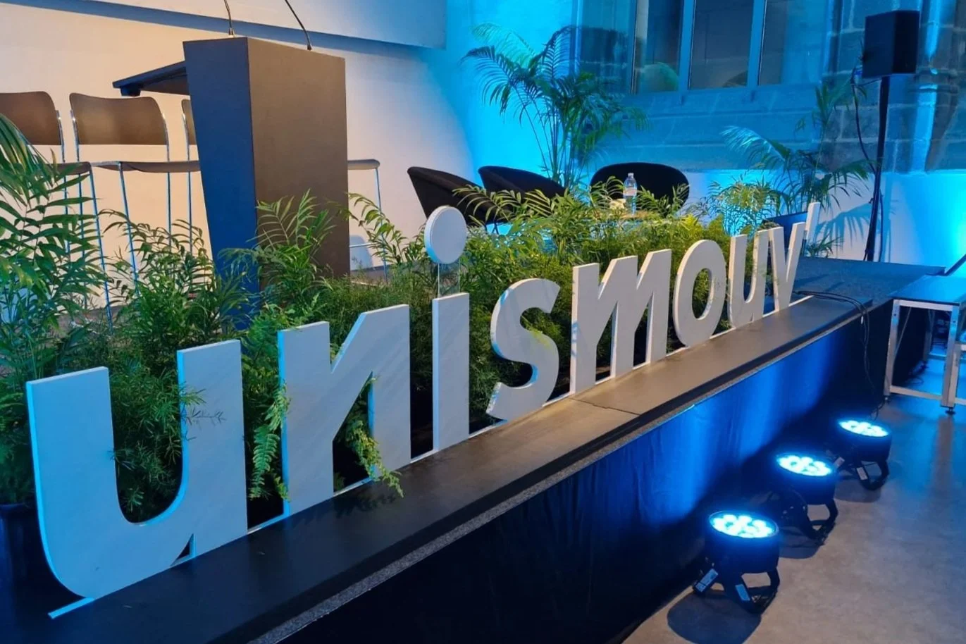 Une scène d'événement avec une grande enseigne lumineuse en lettres blanches formant le mot 'UNISSMOUV'. La scène est décorée avec des plantes vertes, un fond éclairé en bleu, et quelques chaises et tables visibles en arrière-plan.