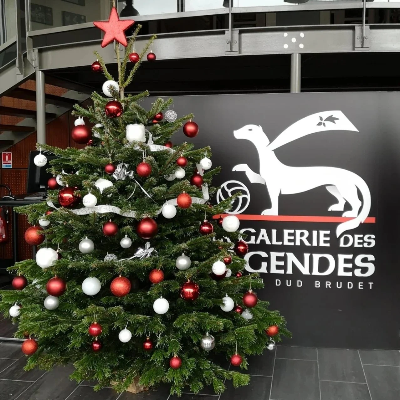 Sapin de Noël décoré de boules rouges, blanches, et argentées, avec une étoile rouge au sommet, placé devant un mur noir avec un logo blanc d'un lézard et le texte 'Galerie des Gendes DUD BRUDET'.