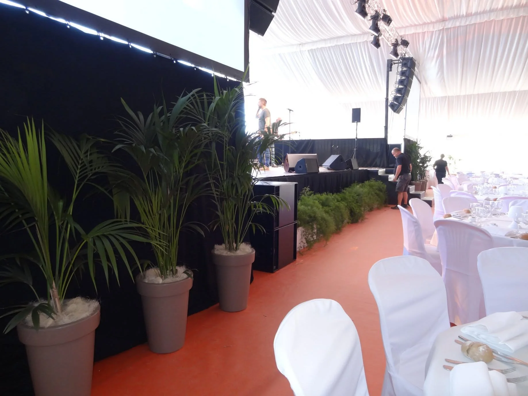 Une scène intérieure d'un événement ou d'une réception avec une scène en arrière-plan, des tables dressées avec des serviettes blanches et des plats, ainsi que des plantes vertes en pots, dans un espace sous une tente ou un grand chapiteau.
