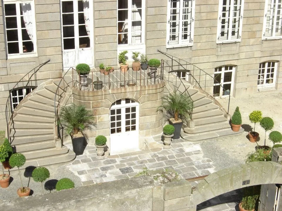 Escalier en pierre menant à une porte d'entrée, entouré de plantes en pot et de balustrades en fer forgé.