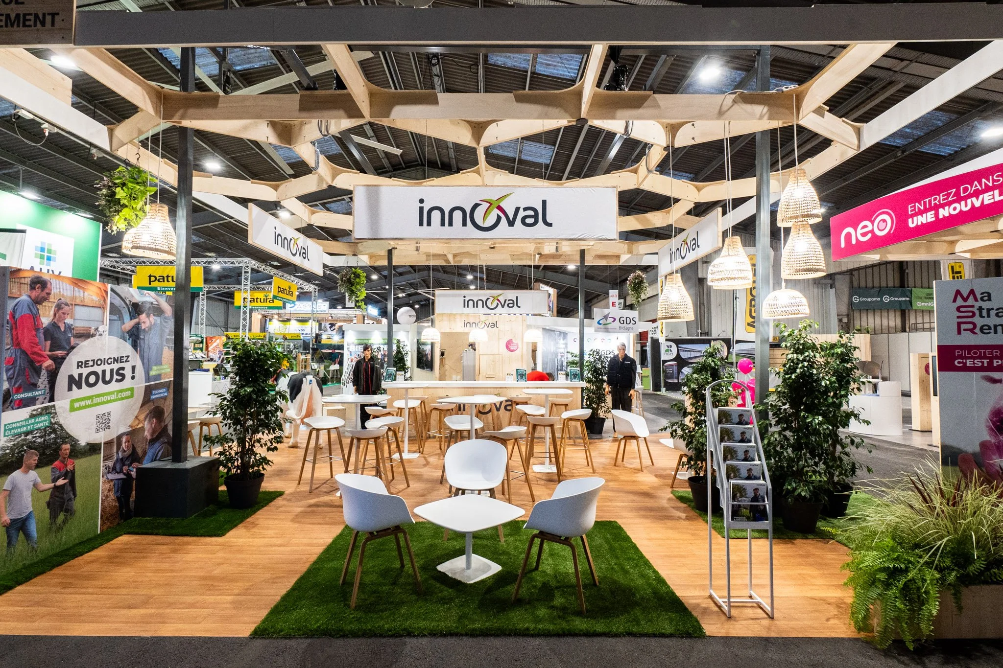 Un stand d'exposition avec des sièges blancs et des tables, décoré avec des plantes, des lampes en osier et un panneau avec le logo 'innoval' suspendu au plafond. Autre stand visible avec des affiches et des panneaux promotionnels, dans un environnement intérieur d'exposition ou de salon professionnel.